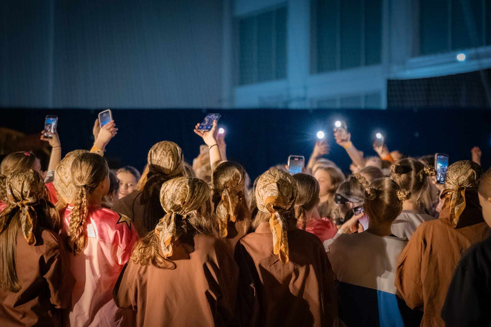 Widok z tyłu na grupę dziewcząt w jednakowych chustach, trzymających telefony z włączonymi lampami błyskowymi podczas wydarzenia.