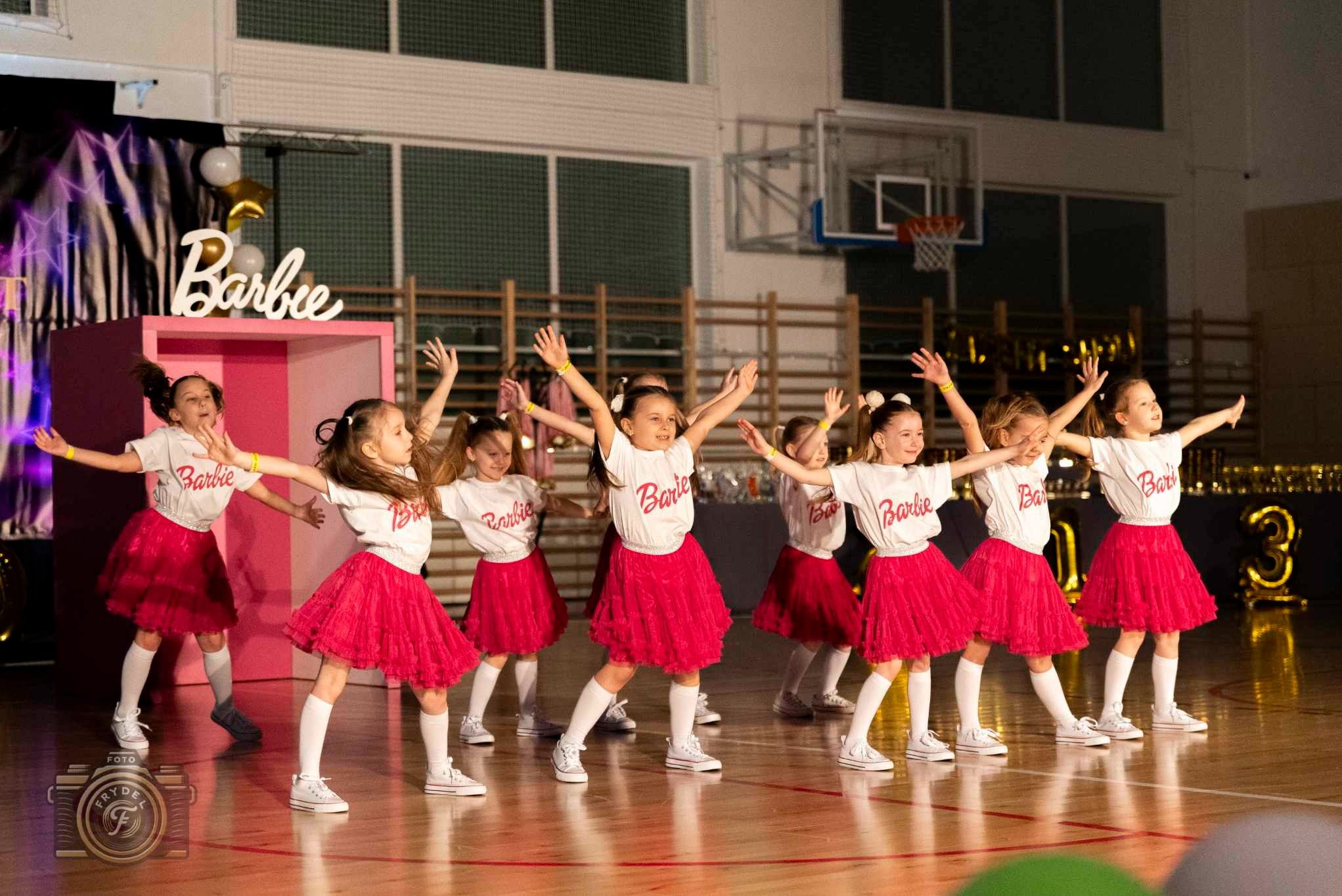 Grupa dziewcząt w strojach Barbie tańczy na scenie. Ubrane w białe koszulki i czerwone spódniczki, z podniesionymi rękami, w tle dekoracja z napisem Barbie.