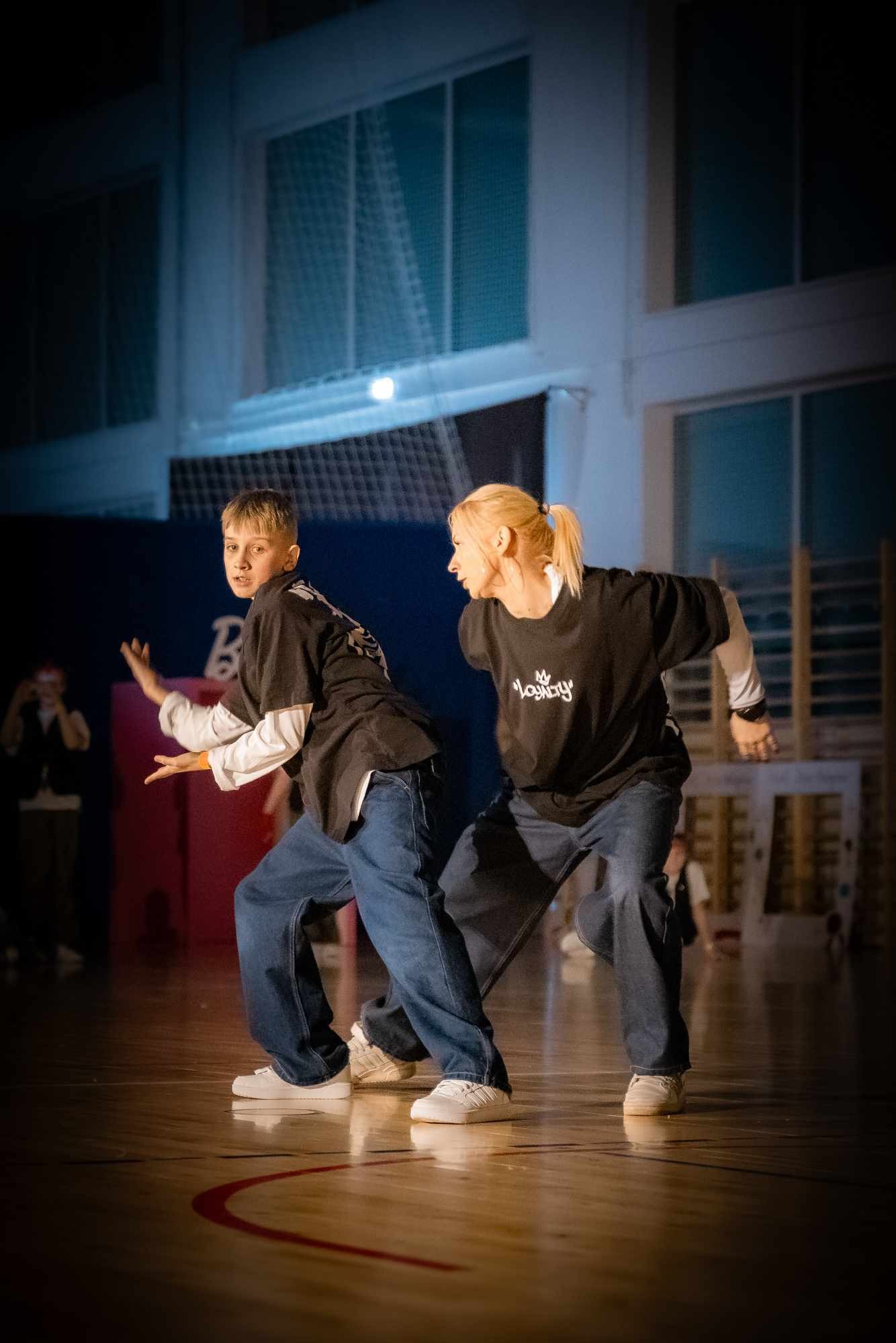 Dynamiczne ujęcie duetu tanecznego w trakcie występu na parkiecie. Młody chłopak i kobieta w luźnych strojach prezentują zsynchronizowane ruchy w hali sportowej.