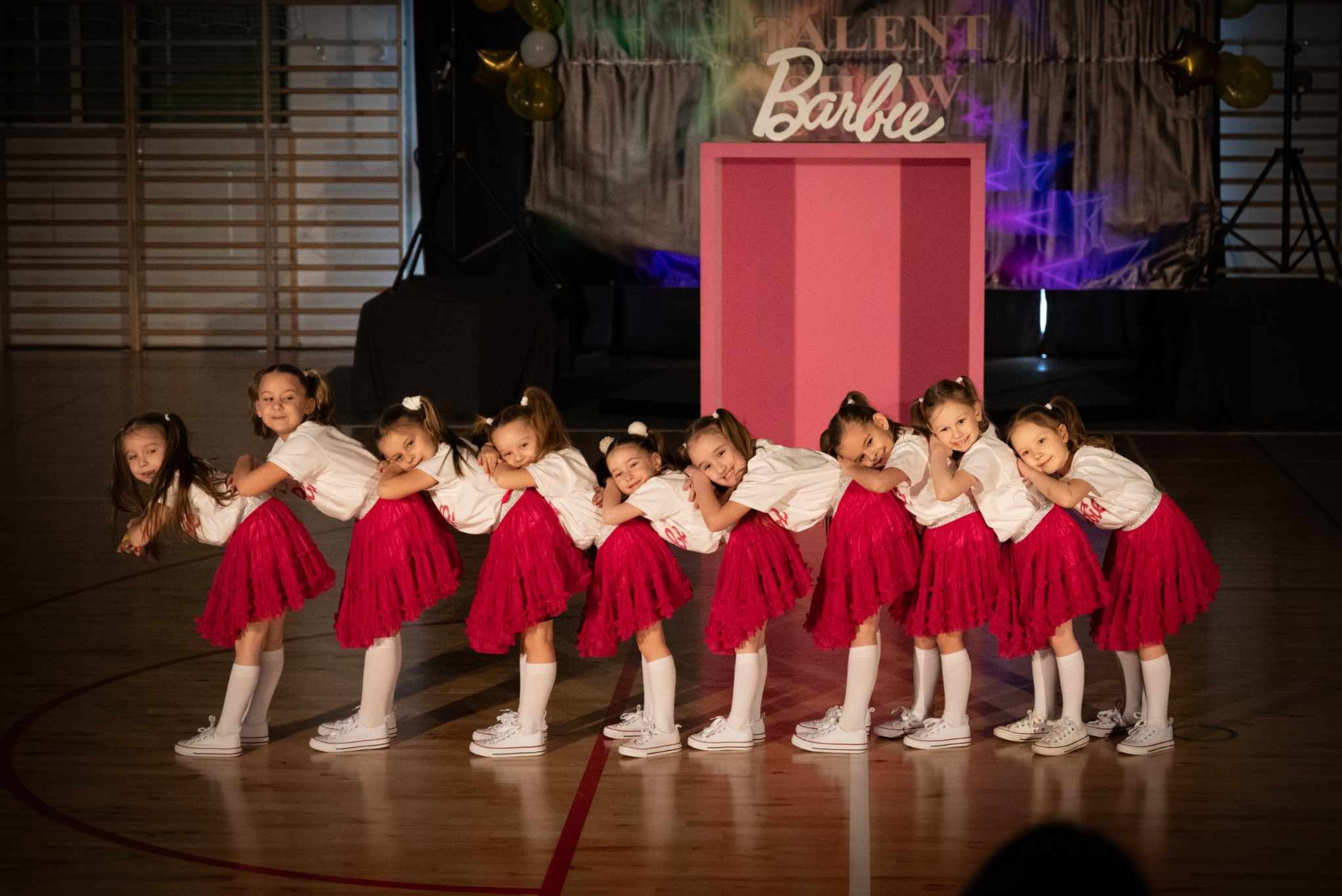 Grupa dziewczynek w białych koszulkach i czerwonych spódniczkach pozuje na tle sceny z dekoracjami 'Talent Barbie'. Uśmiechnięte twarze, taneczny układ.