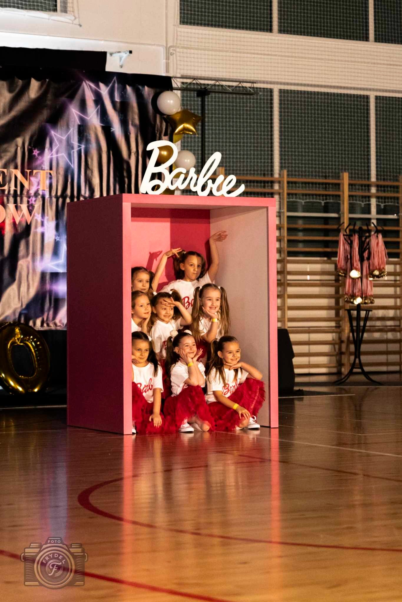 Grupowe zdjęcie dziewczynek w stylizacji Barbie, w różowej budce na tle sali gimnastycznej z dekoracjami eventowymi i medalami. Uśmiechnięte dzieci w białych koszulkach i czerwonych spódniczkach.