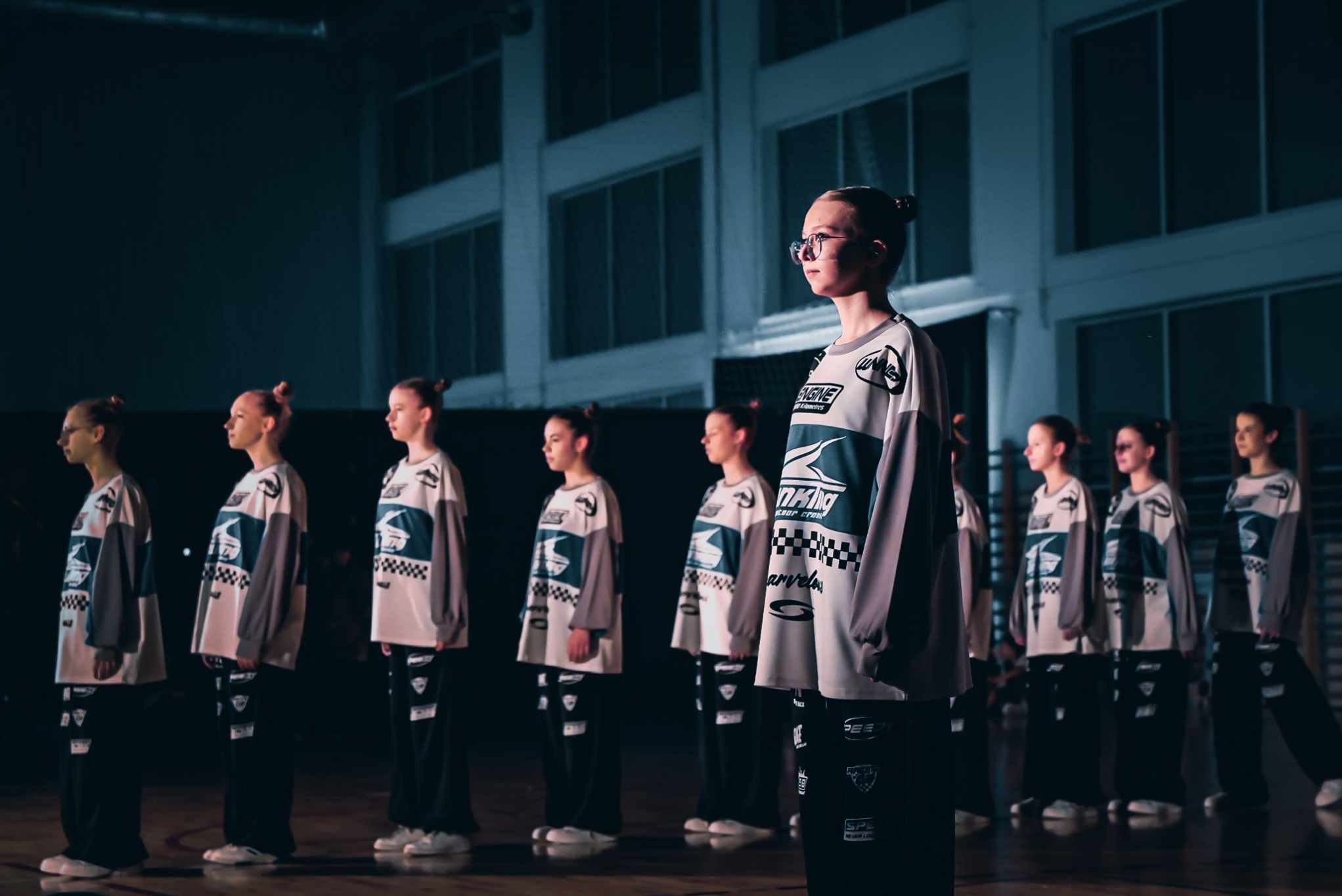 Grupa tancerek w sportowych strojach ustawiona w rzędzie na sali gimnastycznej, oświetlona punktowym światłem, z przodu dziewczyna w okularach.