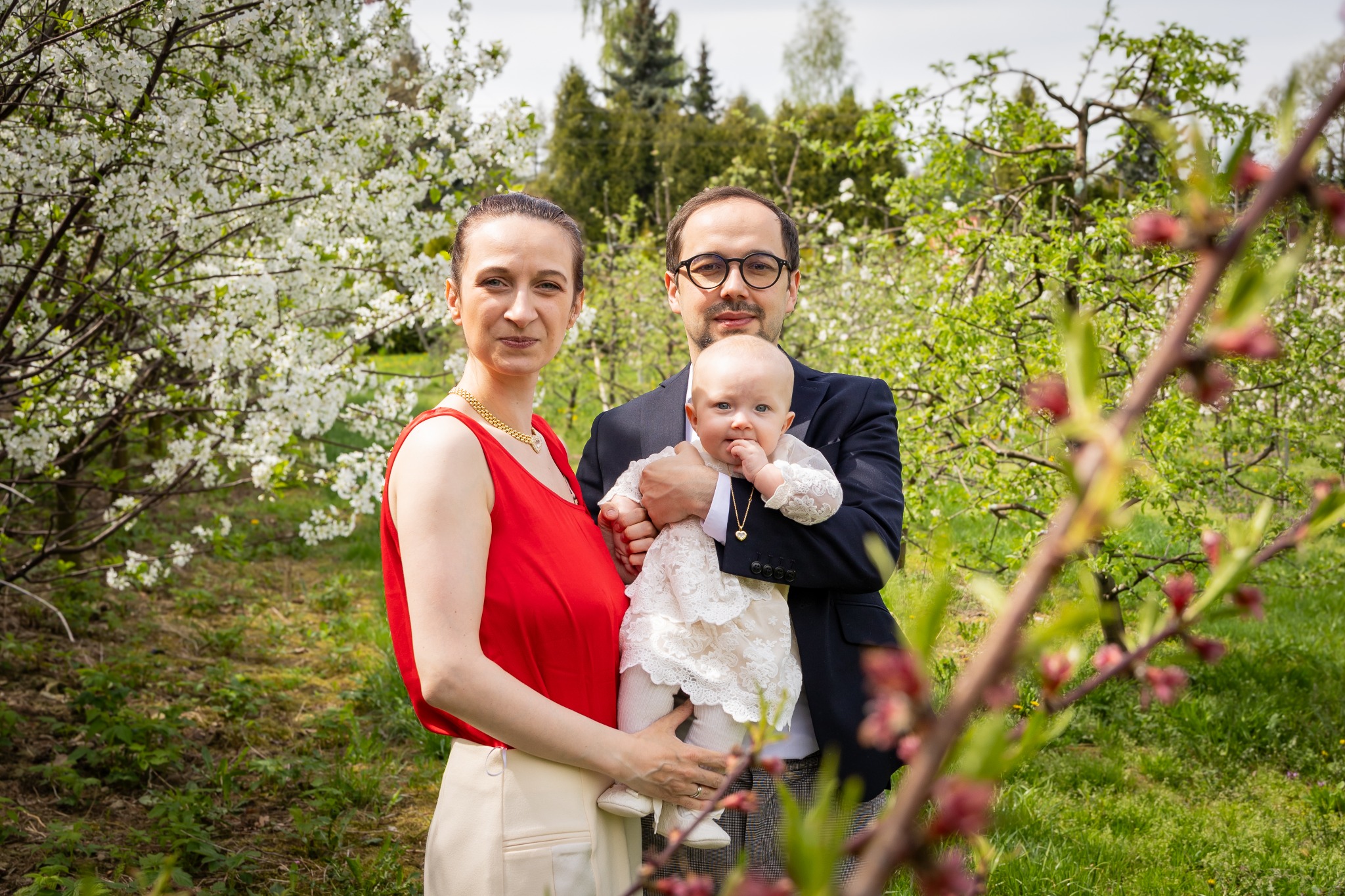 Rodzinne zdjęcie w plenerze: mama w czerwonej bluzce i tata w marynarce trzymają ubraną w białą sukienkę córeczkę na tle kwitnących drzew.