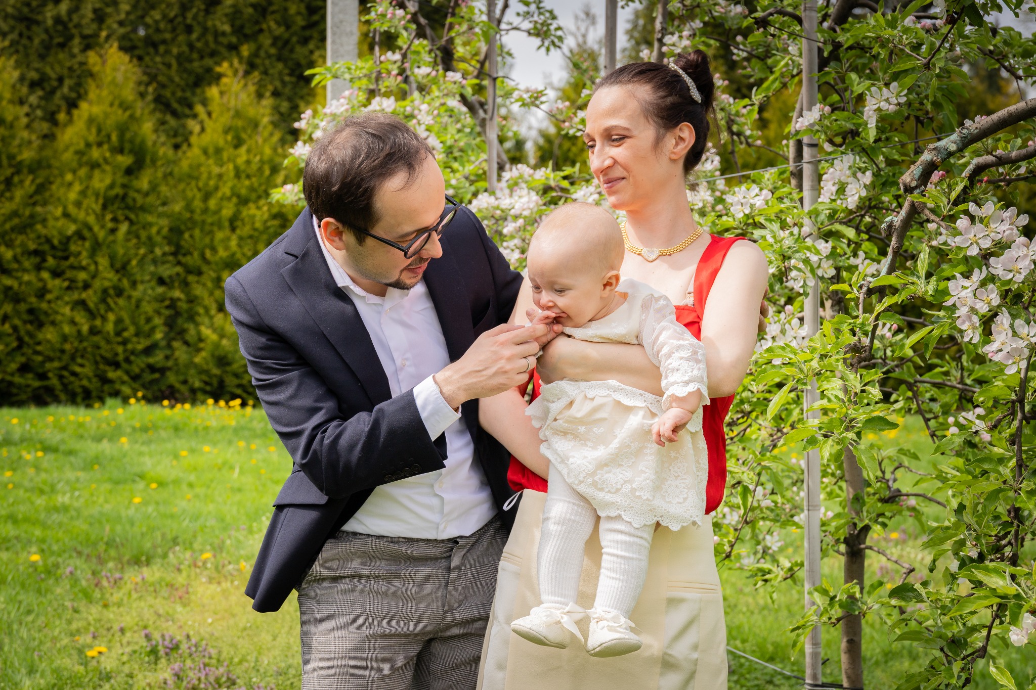 Uśmiechnięta rodzina z niemowlęciem w kwitnącym sadzie. Dziecko w białej sukience, rodzice w eleganckich strojach. Naturalne światło, wiosenna sceneria.