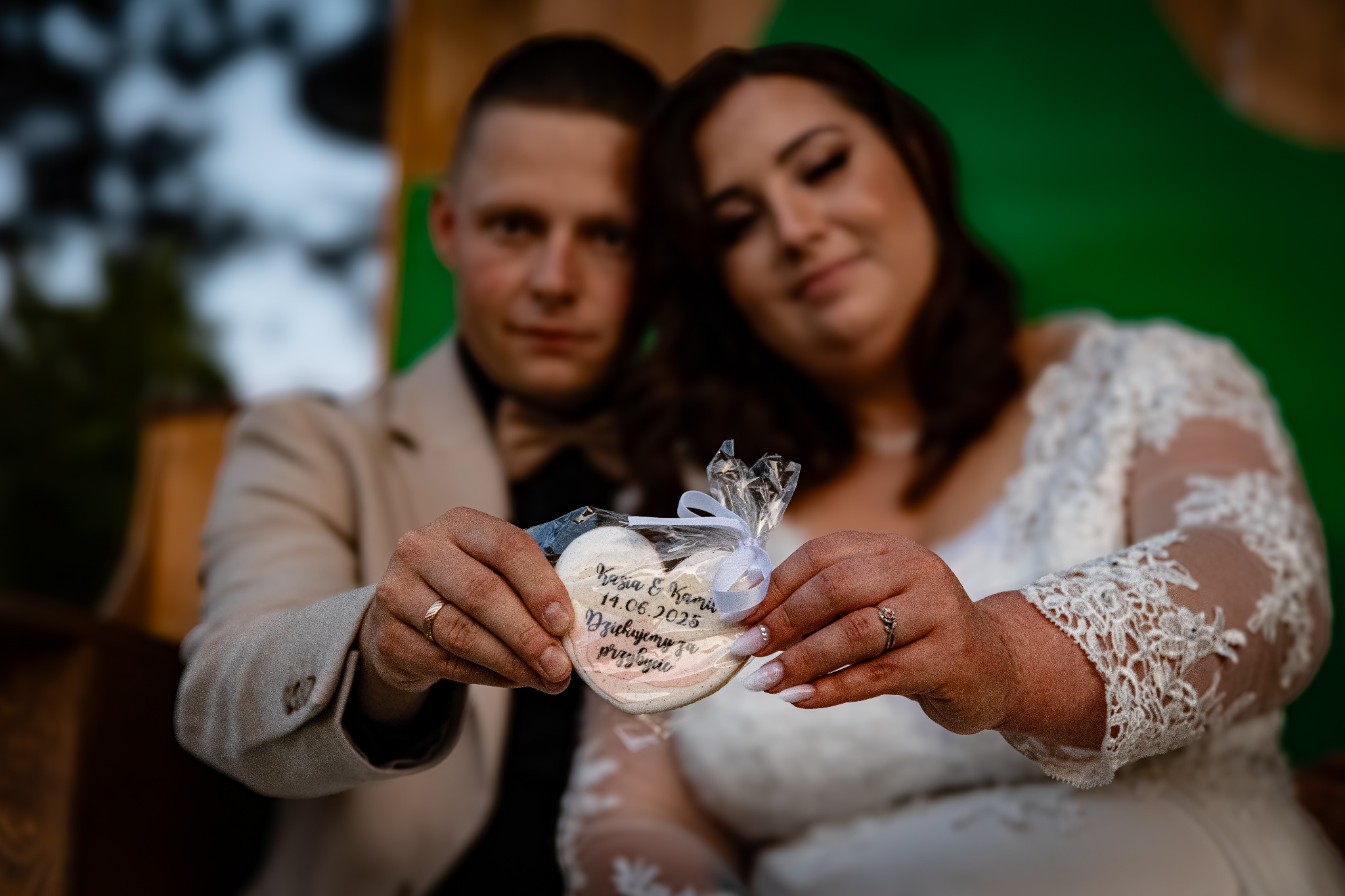 Młoda para z Andrychowa trzyma piernikowe serce z datą ślubu. Panna młoda w koronkowej sukni, pan młody w beżowym garniturze. Ujęcie z bliska, skupione na detalu.