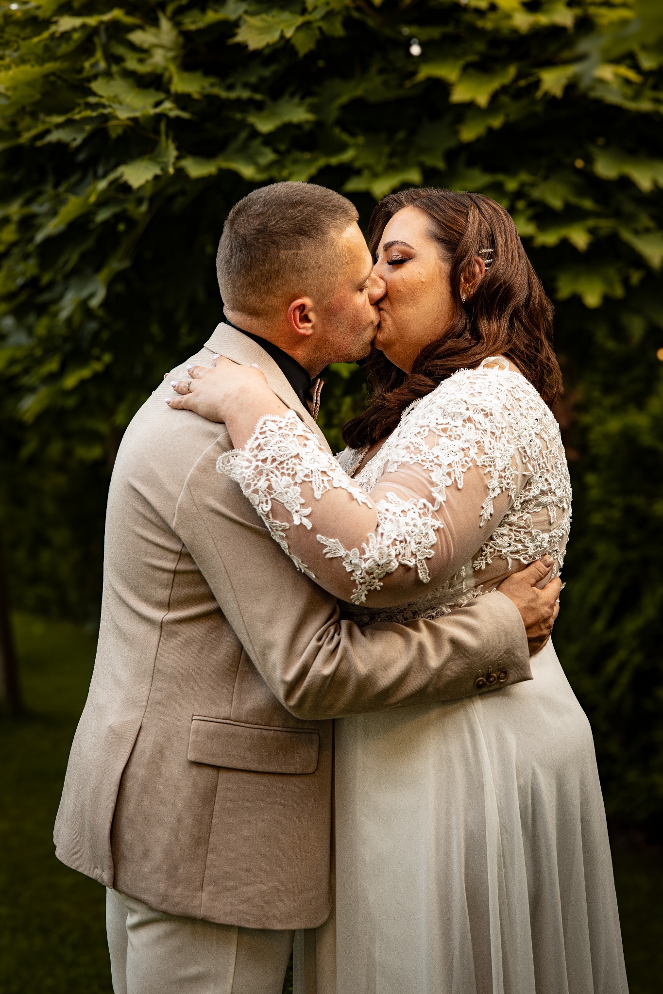Pocałunek pary młodej na tle zieleni: on w beżowym garniturze, ona w koronkowej sukni ślubnej z długimi rękawami. Ujęcie z bliska, skupione na intymnym momencie.