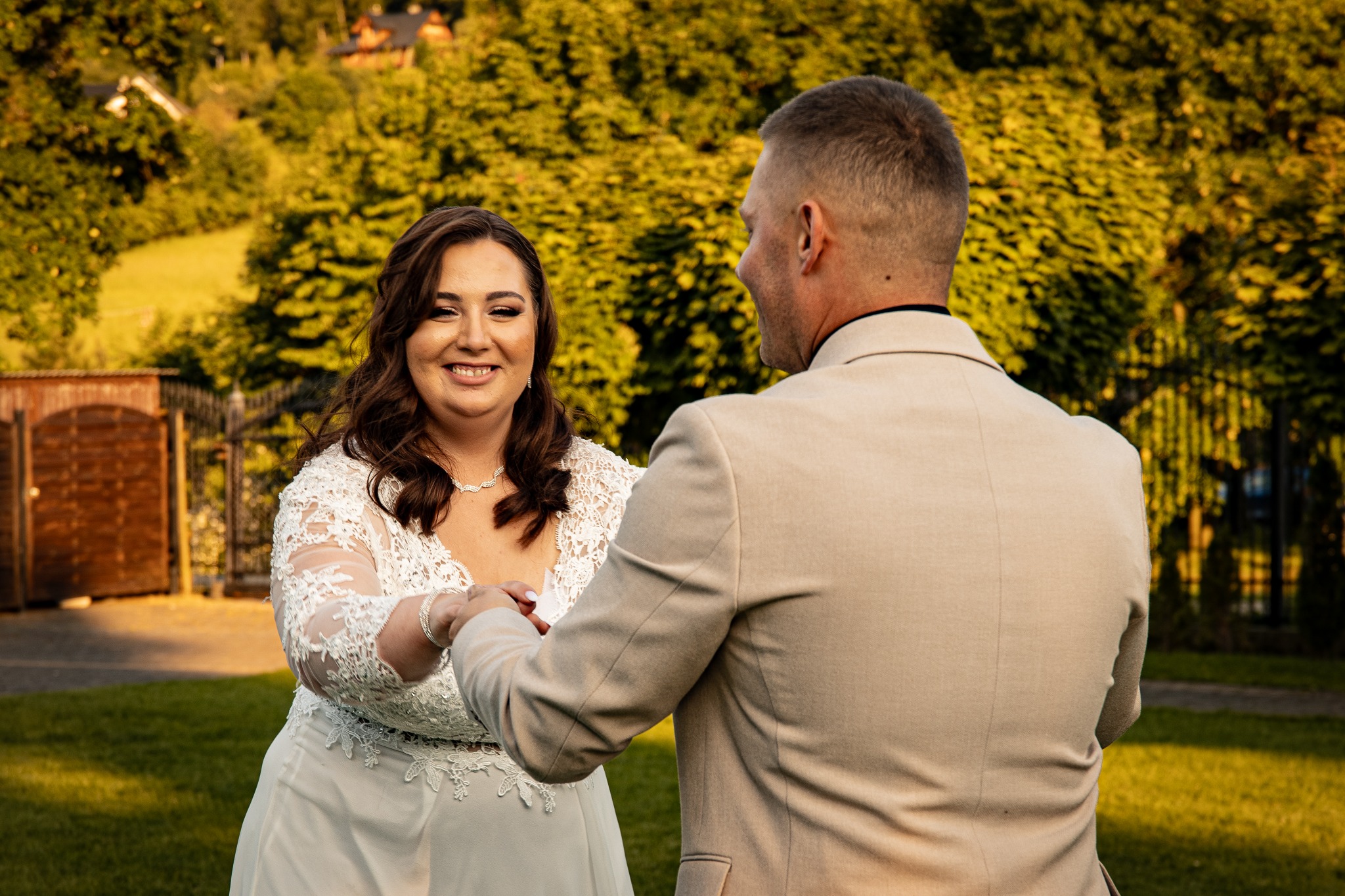 Uśmiechnięta panna młoda w koronkowej sukni trzyma za rękę pana młodego na tle zieleni i drewnianej architektury.