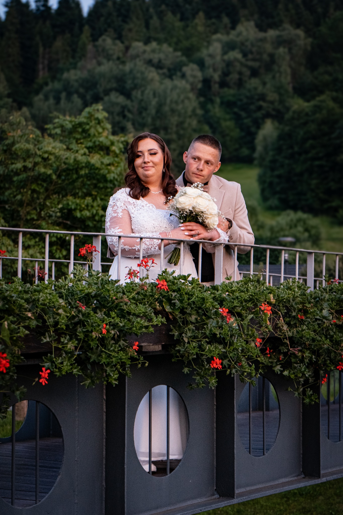 Para młoda w zwiewnej sukni i beżowym garniturze pozuje z bukietem białych róż na tle zieleni, opierając się o balustradę z czerwonymi kwiatami.