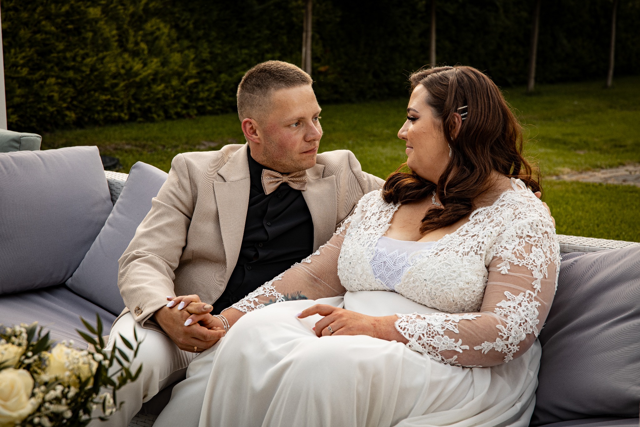 Para młoda siedzi na sofie, trzymając się za ręce. Pan młody w beżowym garniturze i muszce, panna młoda w białej sukni z koronkowymi rękawami. W tle zieleń.
