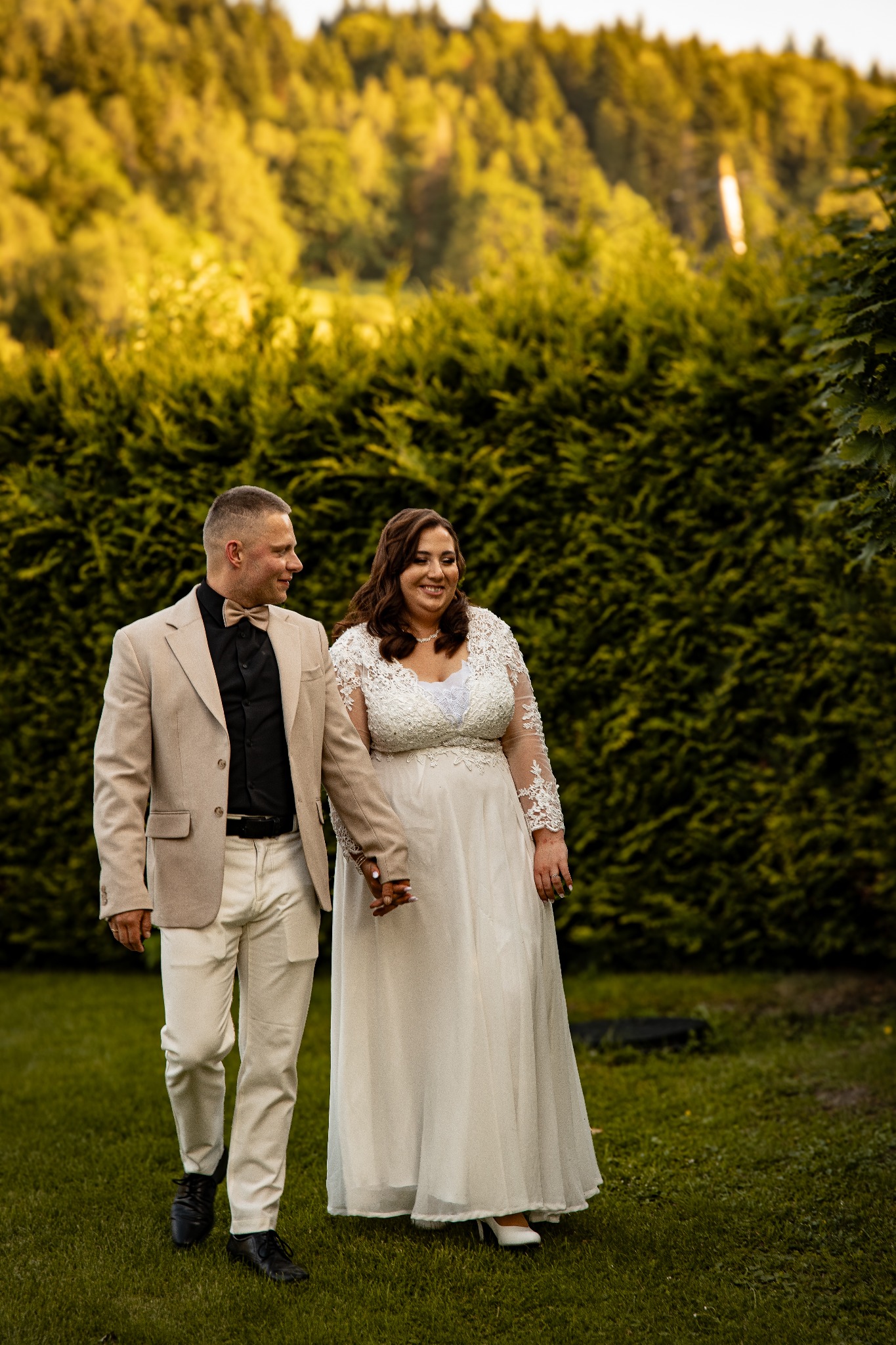 Uśmiechnięta para młoda, trzymając się za ręce, spaceruje po zielonym trawniku na tle wysokiego, zielonego żywopłotu w słoneczny dzień.