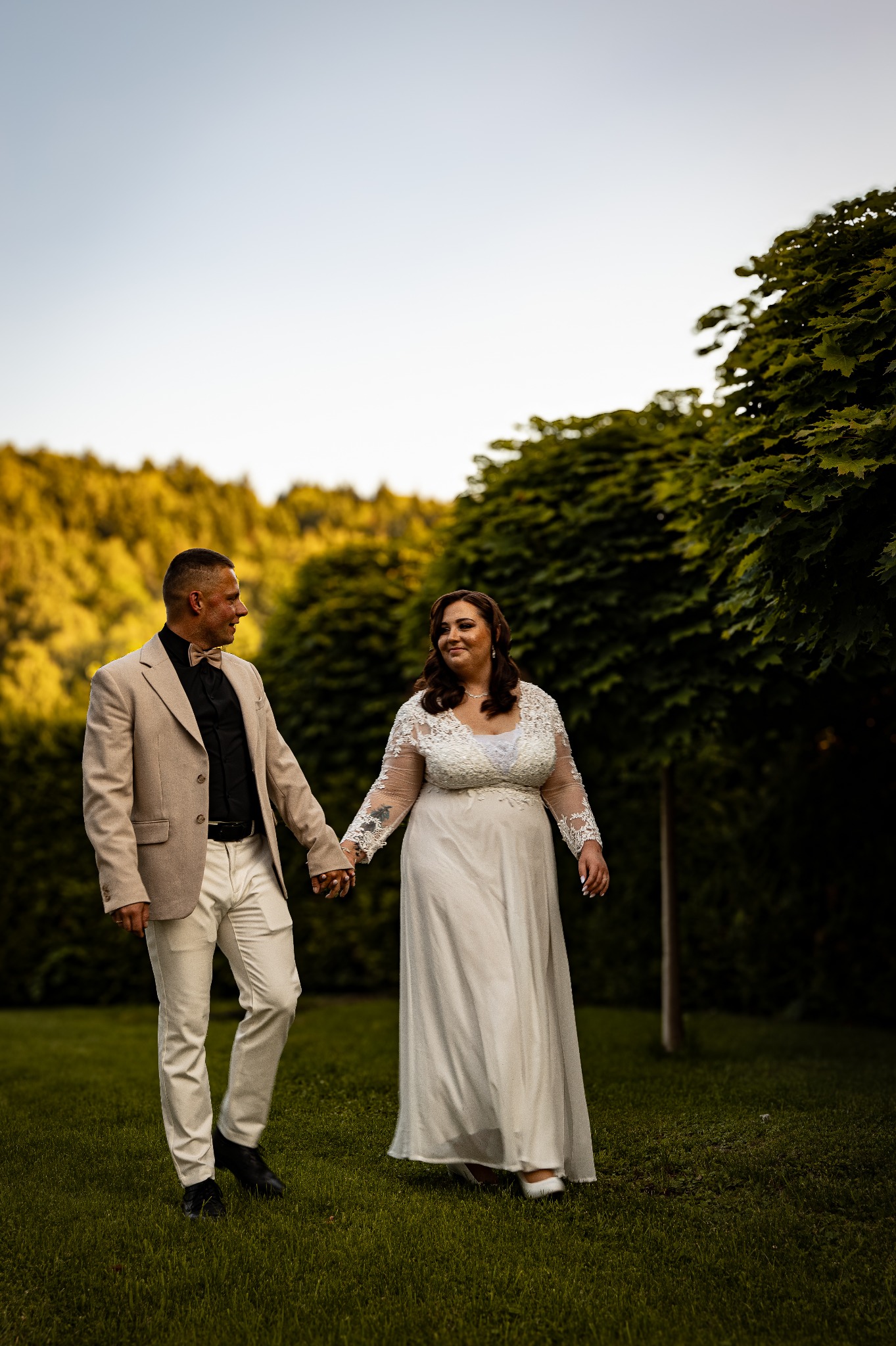 Para młoda, trzymając się za ręce, spaceruje po zielonym trawniku w plenerze. Pan młody w beżowym garniturze, panna młoda w białej sukni z koronkowymi rękawami.