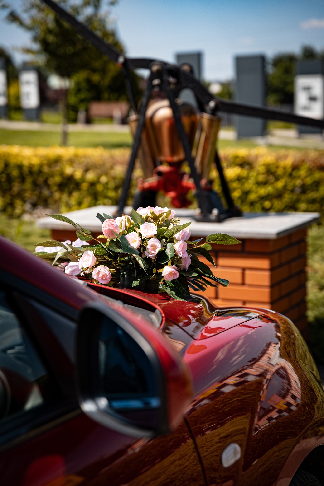 Bukiet różowych róż na karoserii czerwonego auta, w tle abstrakcyjna rzeźba i zieleń parku w słoneczny dzień. Dekoracja ślubna na samochodzie.