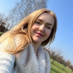 Dialogi Terapeutyczne - Uśmiechnięta blond kobieta w futrzanym płaszczu, robiąca selfie na tle parku z drzewami i błękitnym niebem.