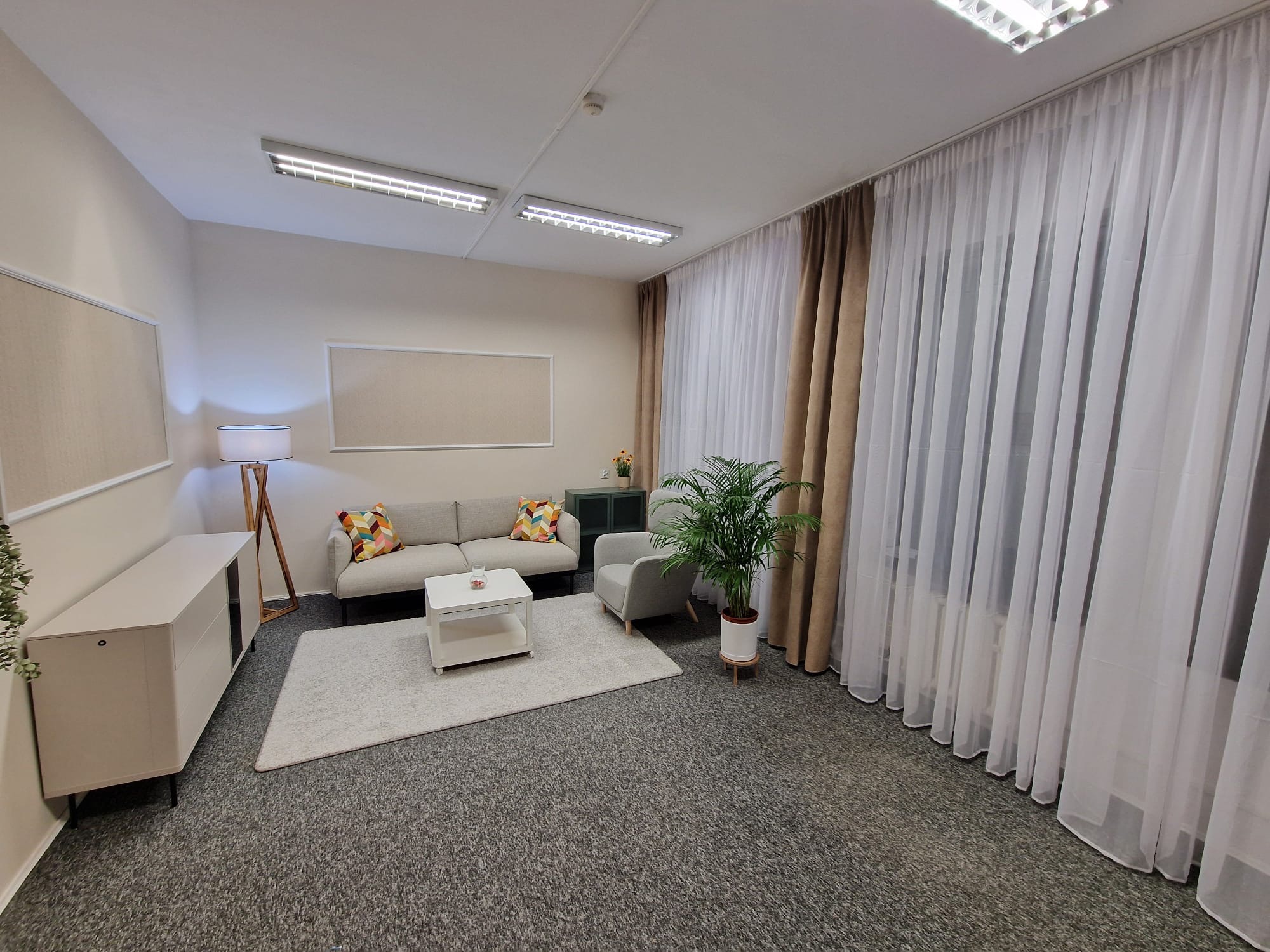 Jasne, przestronne wnętrze gabinetu z sofą, fotelem, stolikiem kawowym i roślinami doniczkowymi, oświetlone lampami sufitowymi i lampą podłogową, z zasłonami w oknach.