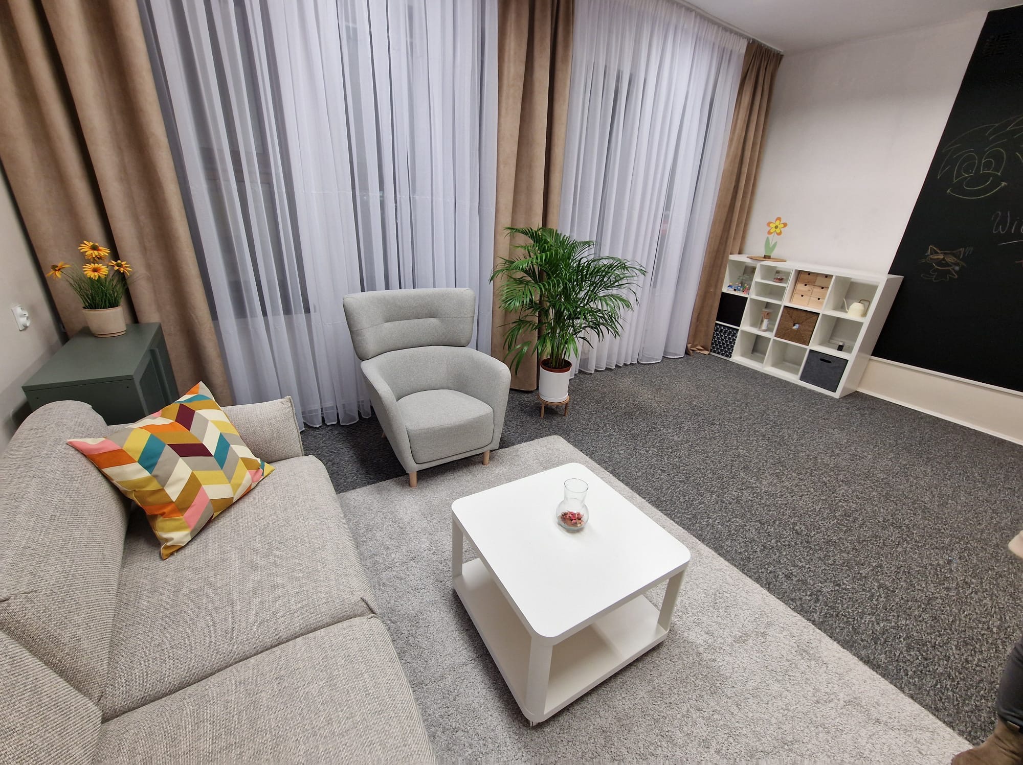 Jasne, przestronne wnętrze gabinetu z szarą sofą z kolorową poduszką, fotelem, białym stolikiem kawowym z wazonem, kwiatami w doniczkach i tablicą z rysunkami.