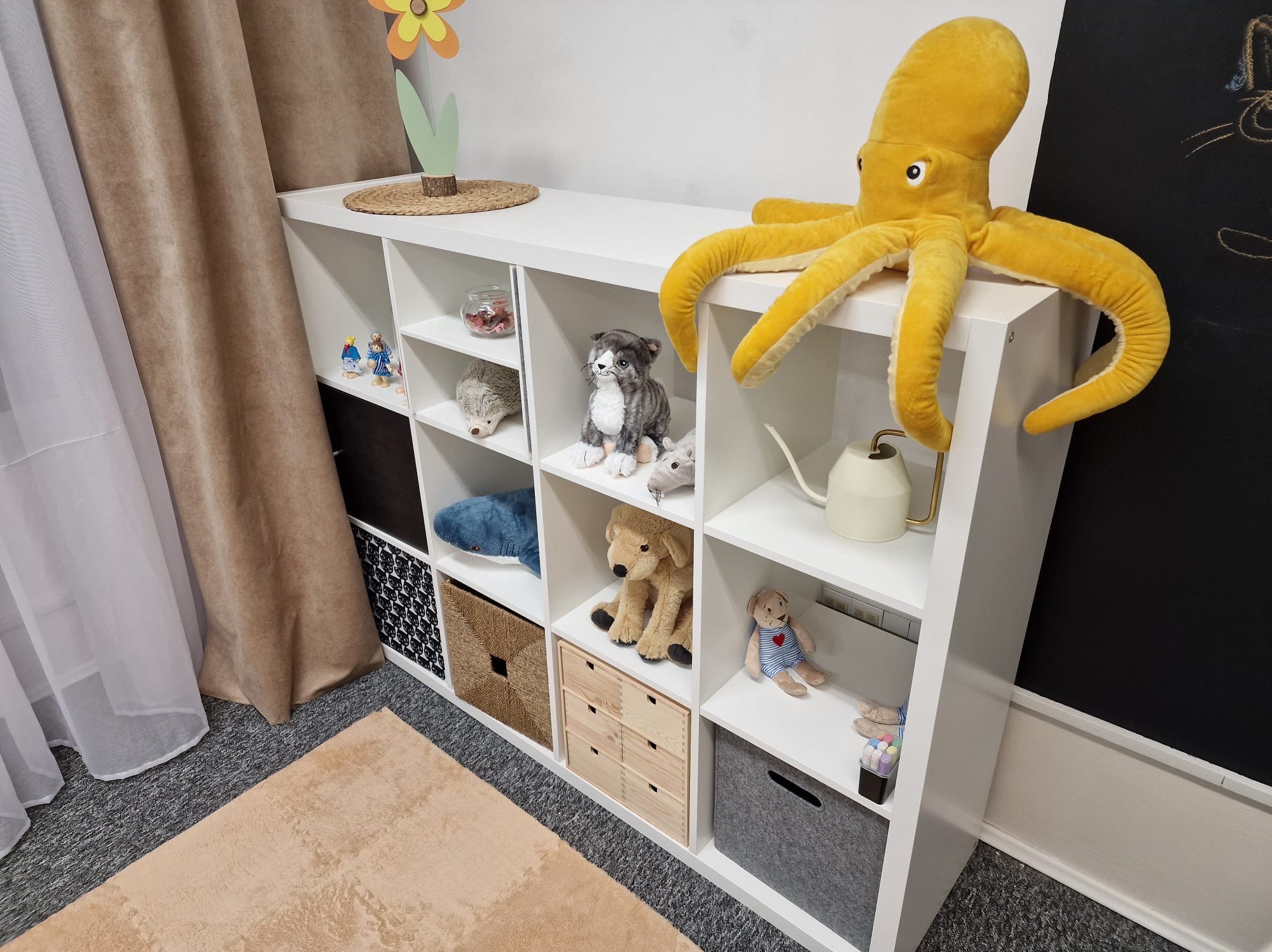 Pokój dziecięcy w Gorzowie Wielkopolskim z białym regałem wypełnionym pluszakami: ośmiornicą, psem, kotem i jeżem, oraz akcesoriami dla dzieci, obok czarnej tablicy do rysowania kredą.