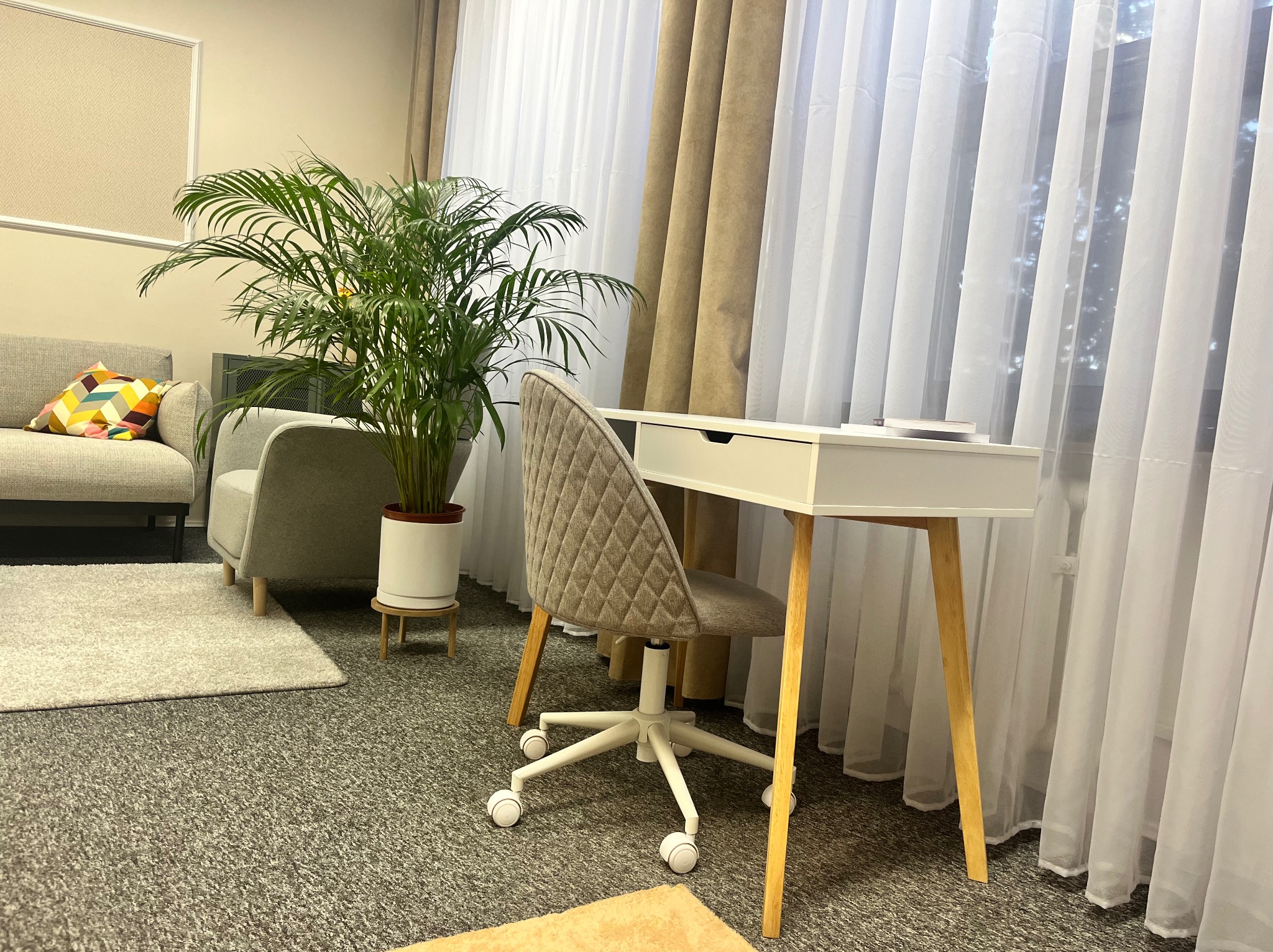 Jasny gabinet psychoterapeuty: biurko z krzesłem na kółkach, roślina doniczkowa, kanapa z kolorową poduszką, zasłony w oknie.