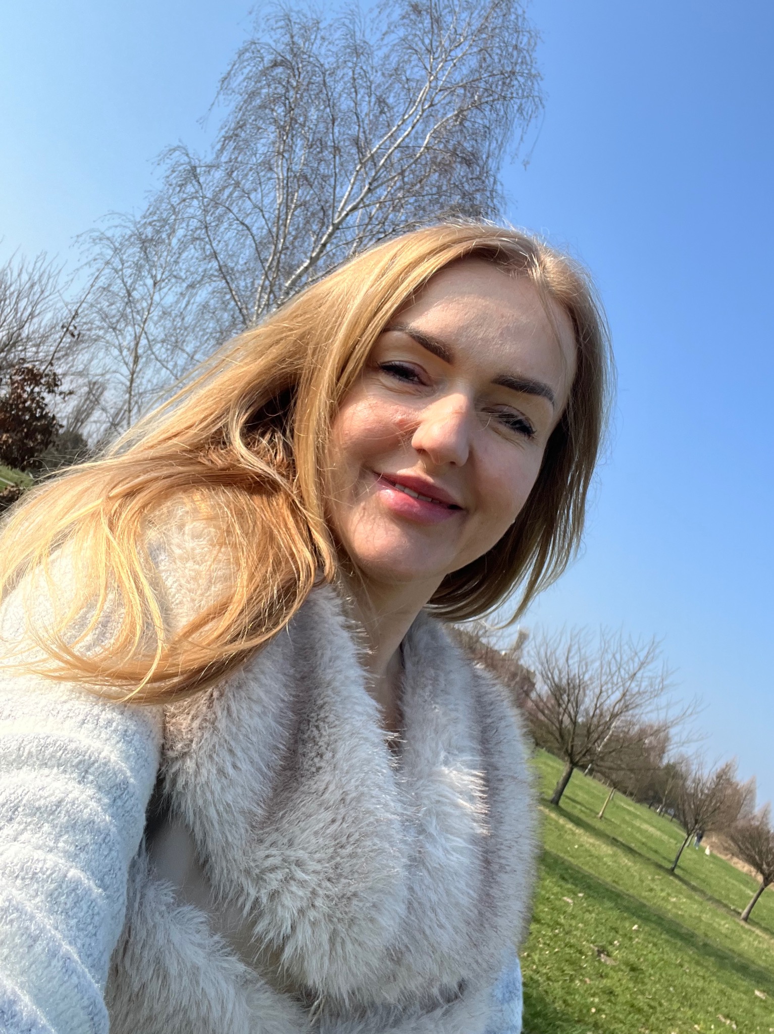 Uśmiechnięta blond kobieta w futrzanym płaszczu, robiąca selfie na tle parku z drzewami i błękitnym niebem.