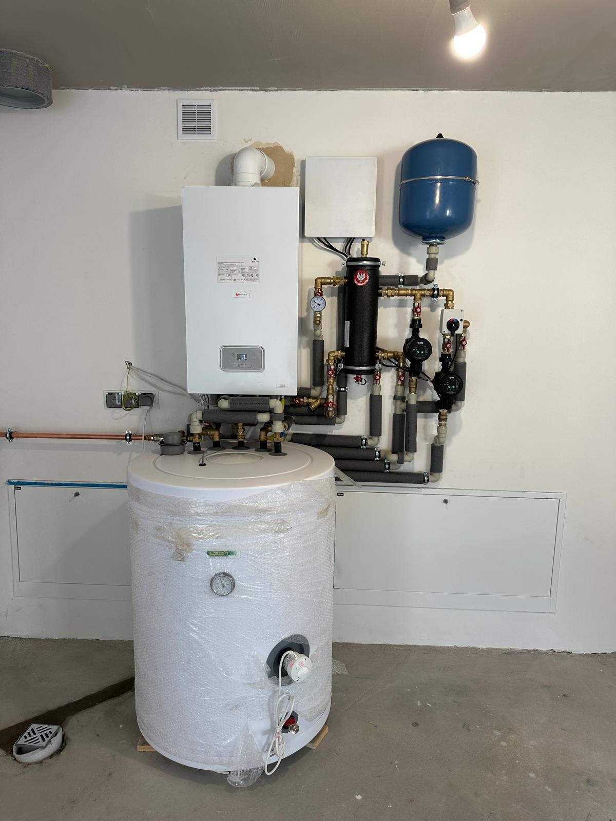 Biały piec CO, zbiornik wody owinięty folią bąbelkową, instalacja z rur miedzianych i zaworów. Całość na tle białej ściany z widoczną wentylacją.