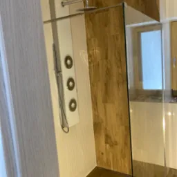 Nowoczesna kabina prysznicowa z panelem natryskowym i drewnopodobnymi płytkami na ścianie, widoczny odpływ liniowy w podłodze.