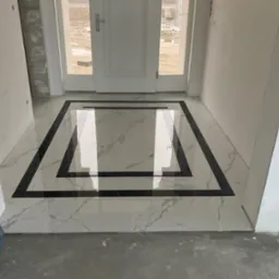 Białe marmurowe płytki podłogowe z czarną geometryczną bordiurą w nowym holu wejściowym, widok na drzwi wejściowe i plac budowy.