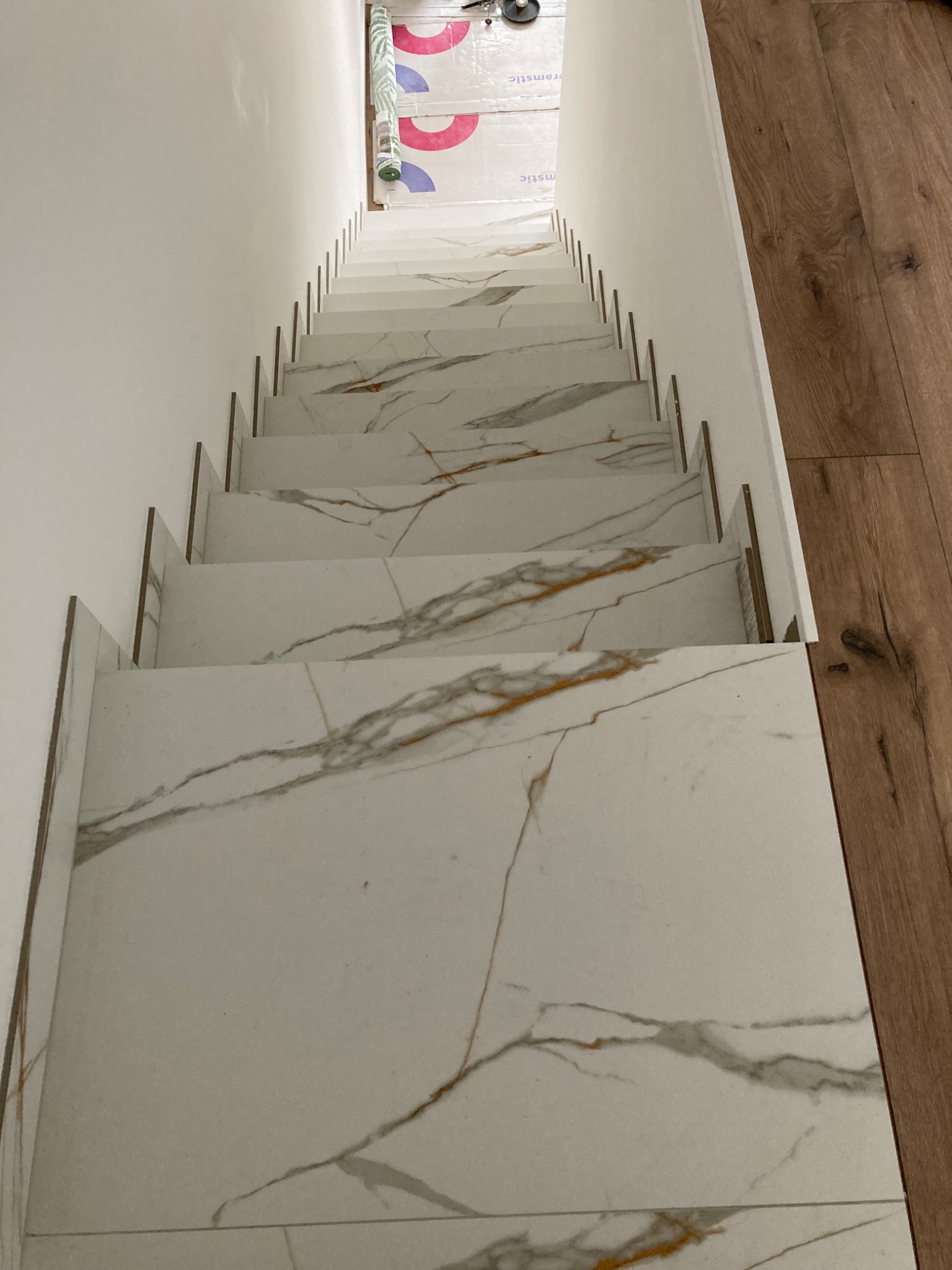 Wykończone schody wewnętrzne z białych płytek imitujących marmur z brązowymi żyłami, widok z góry, na górze schodów folia ochronna.
