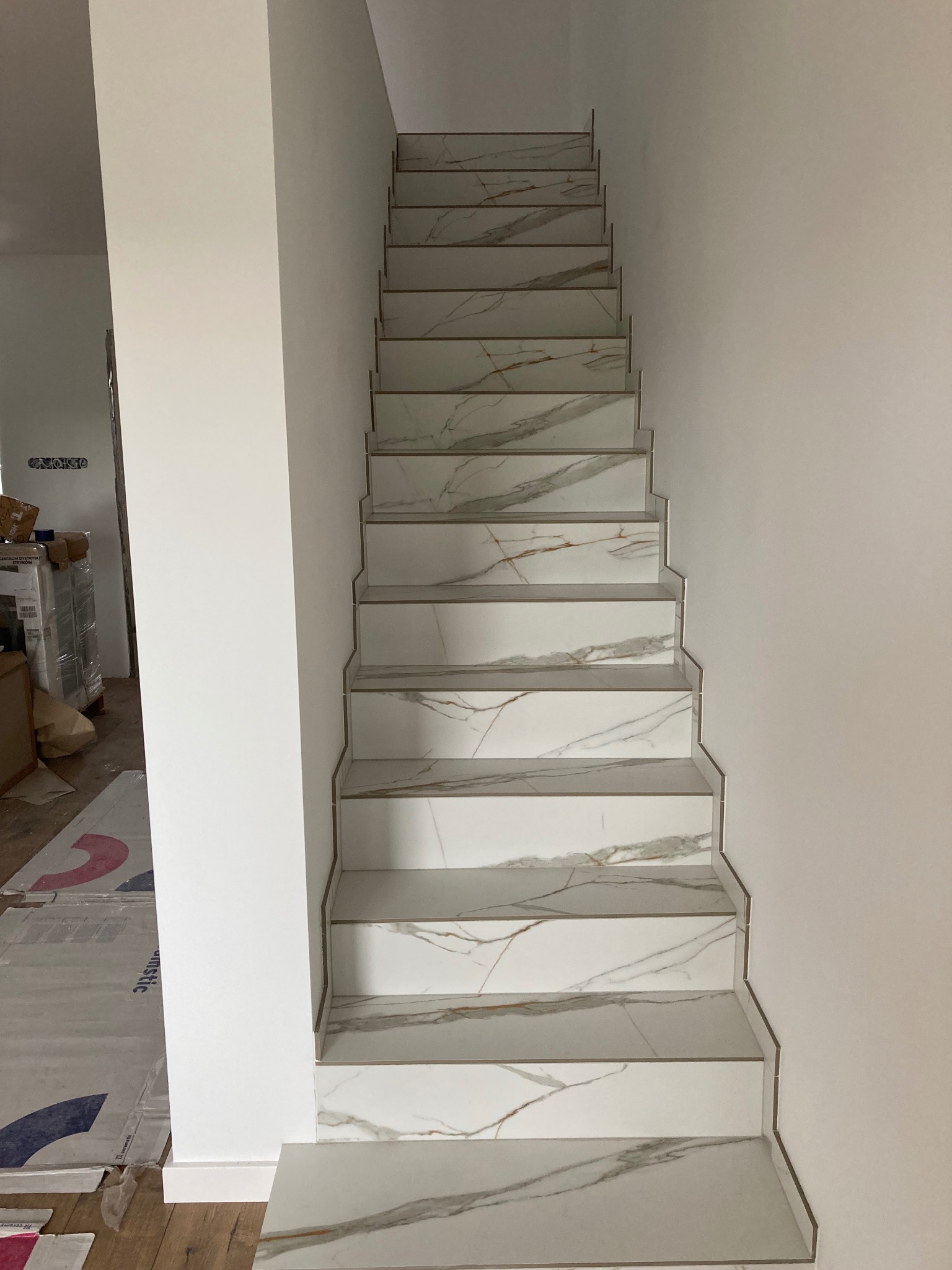 Wykończone schody z białych płytek imitujących marmur z delikatnymi złotymi żyłami, z widocznymi listwami wykończeniowymi, w jasnym pomieszczeniu w trakcie remontu, z zabezpieczoną podłogą.
