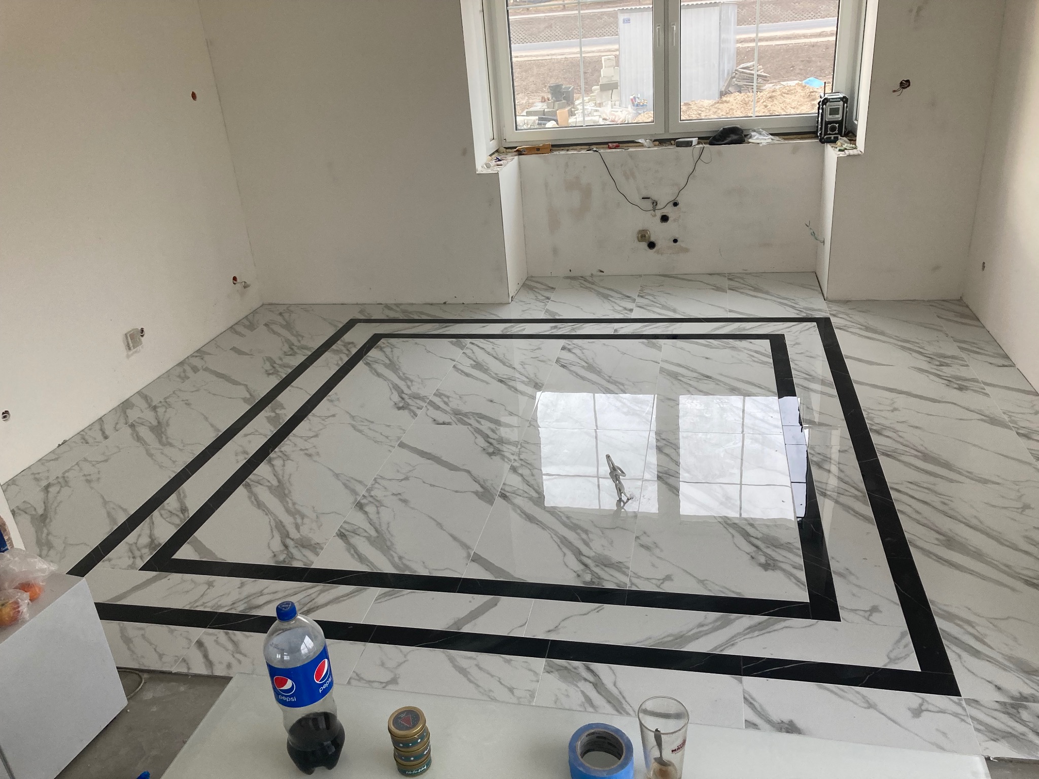 Ułożona podłoga z białych płytek imitujących marmur z czarną ramką, widoczne niedokończone ściany i okno z widokiem na plac budowy