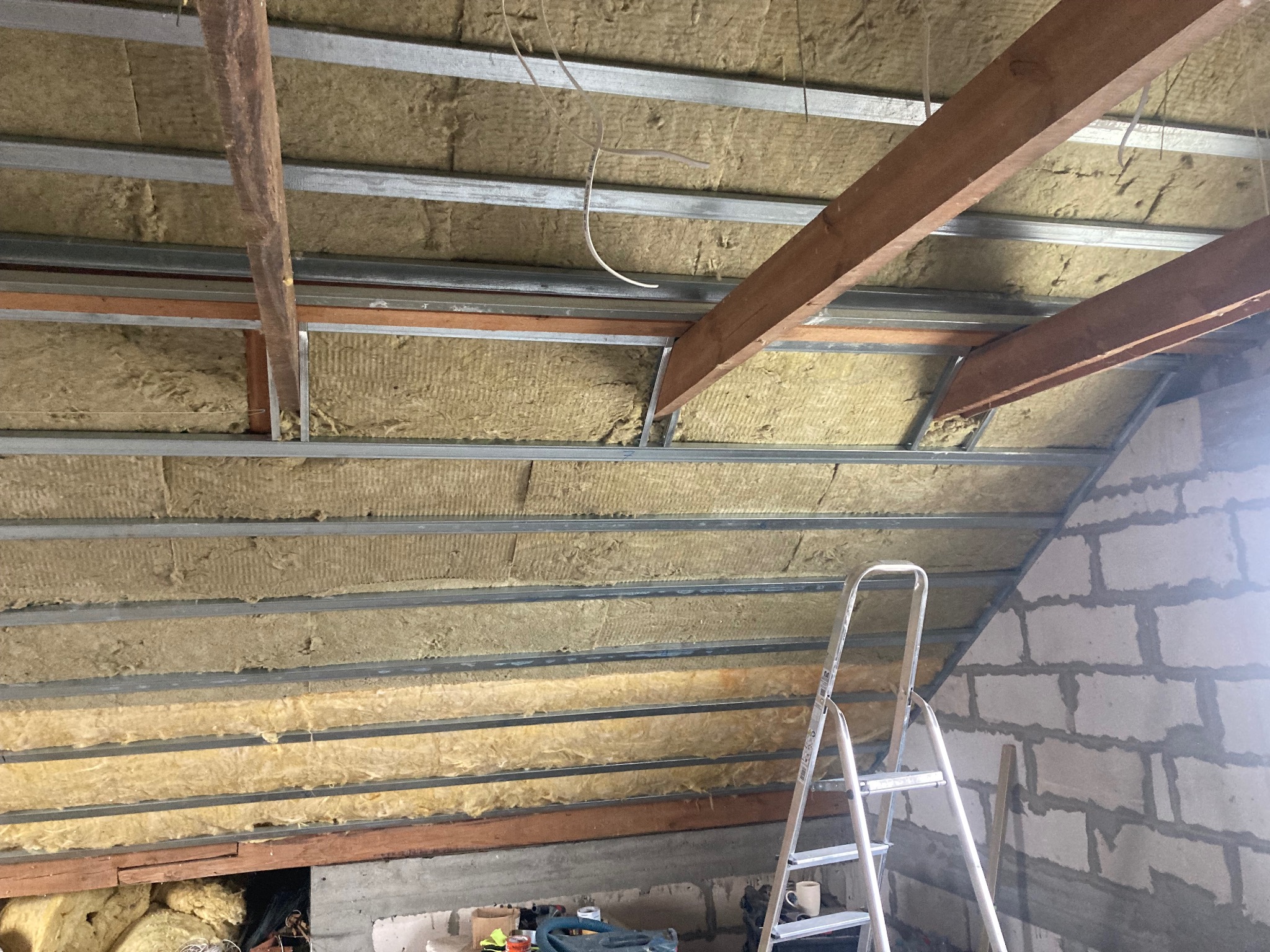 Ocieplenie poddasza wełną mineralną, widoczne metalowe profile, drewniane belki i fragment ściany z bloczków, drabina oparta o ścianę.