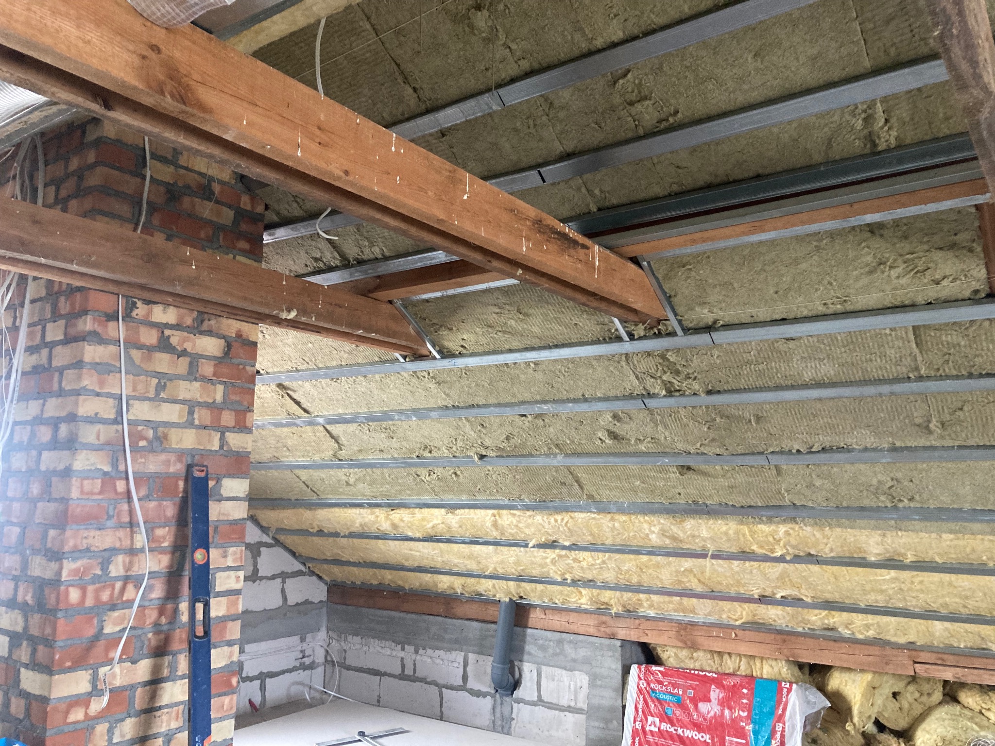 Poddasze w trakcie ocieplania wełną mineralną Rockwool, widoczne drewniane belki, metalowe profile, mur z cegły i poziomica.