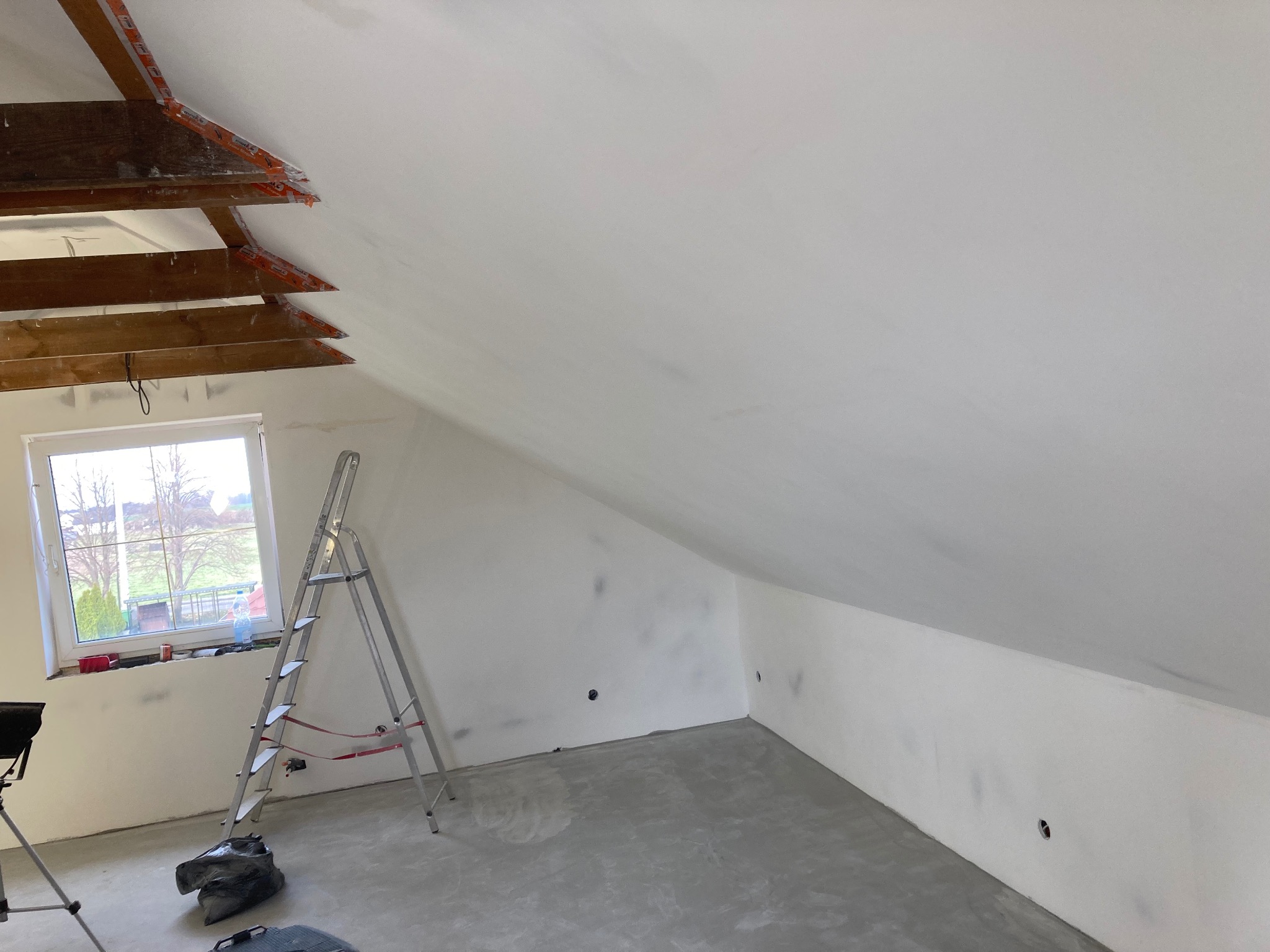 Poddasze po malowaniu na biało, widoczne drewniane elementy konstrukcji dachu zabezpieczone taśmą, drabina oparta o ścianę przy oknie, szpachlowane ściany z widocznymi punktami elektrycznymi.