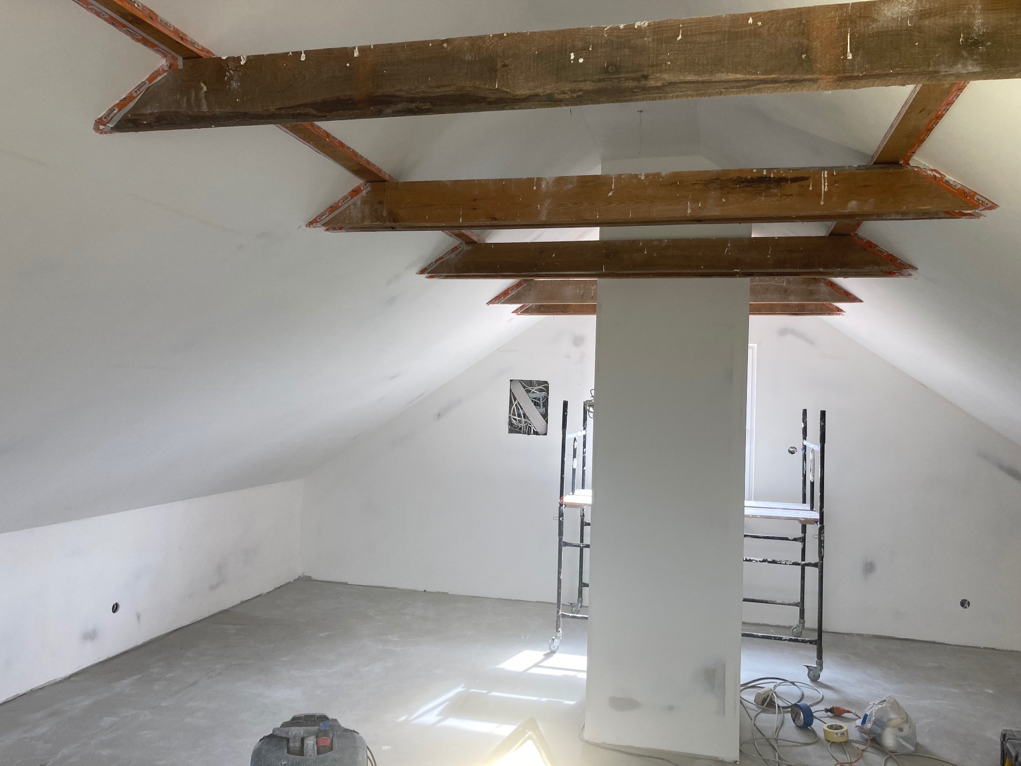 Poddasze w trakcie prac wykończeniowych: surowe drewniane belki stropowe zabezpieczone taśmą malarską, ściany pomalowane na biało, widoczne rusztowania i narzędzia malarskie na podłodze.
