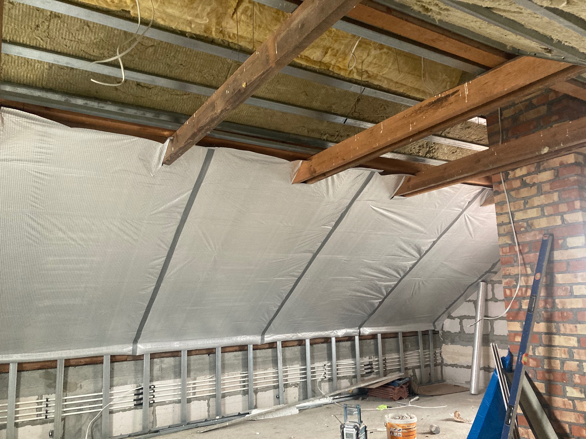 Poddasze w trakcie ocieplania: widoczna izolacja termiczna z folią aluminiową, drewniane belki konstrukcyjne, metalowe profile ścienne, instalacja elektryczna oraz mur z cegły. Poziomica oparta...