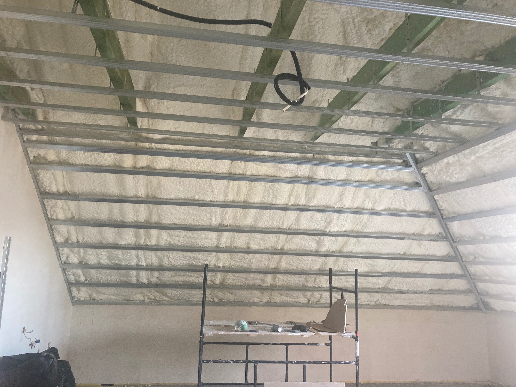Poddasze z metalową konstrukcją i pianką izolacyjną, widoczny fragment rusztowania z narzędziami, przewód elektryczny zwisający z sufitu.