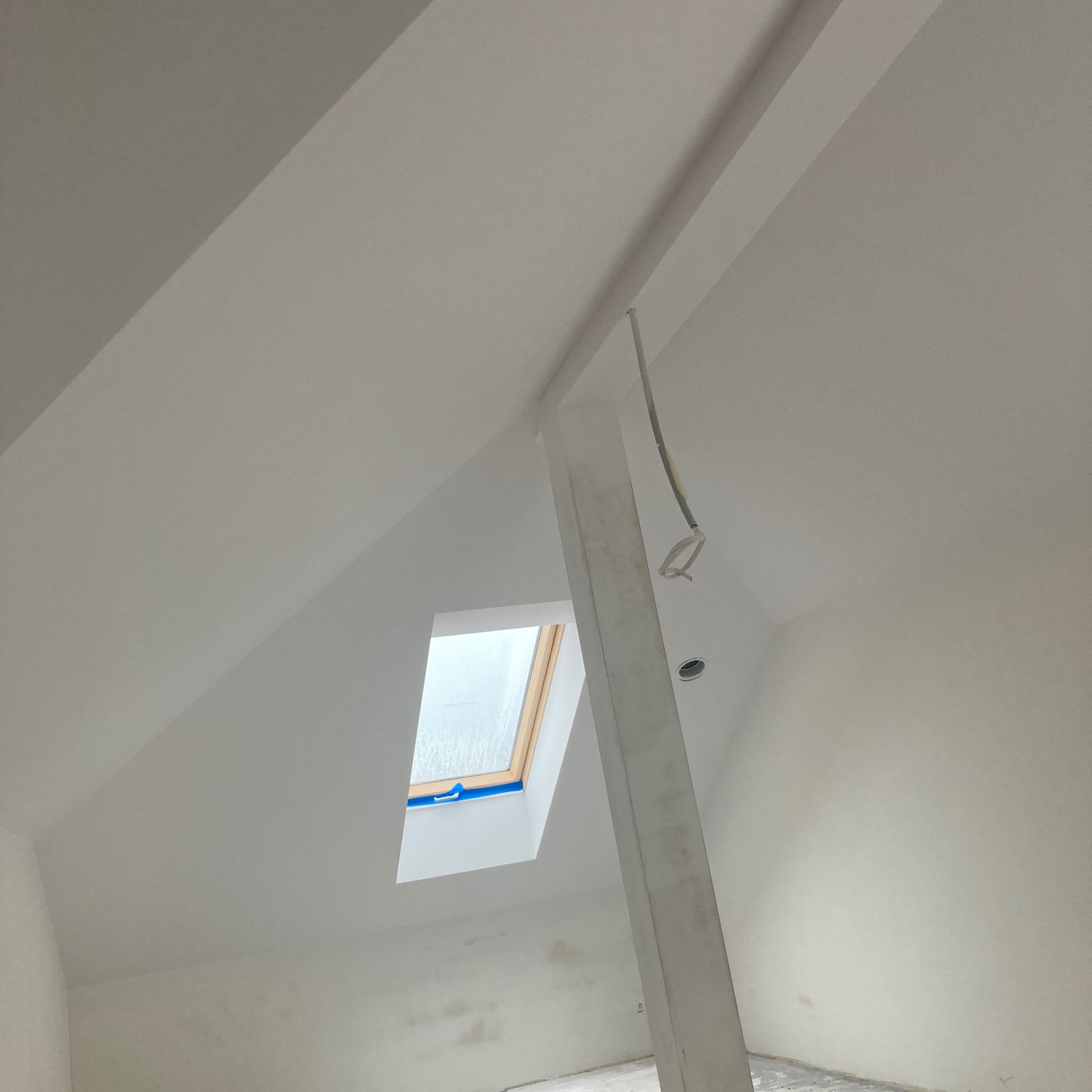 Poddasze po malowaniu na biało, widoczny słup konstrukcyjny, okno dachowe zabezpieczone niebieską taśmą malarską oraz wystające przewody elektryczne.