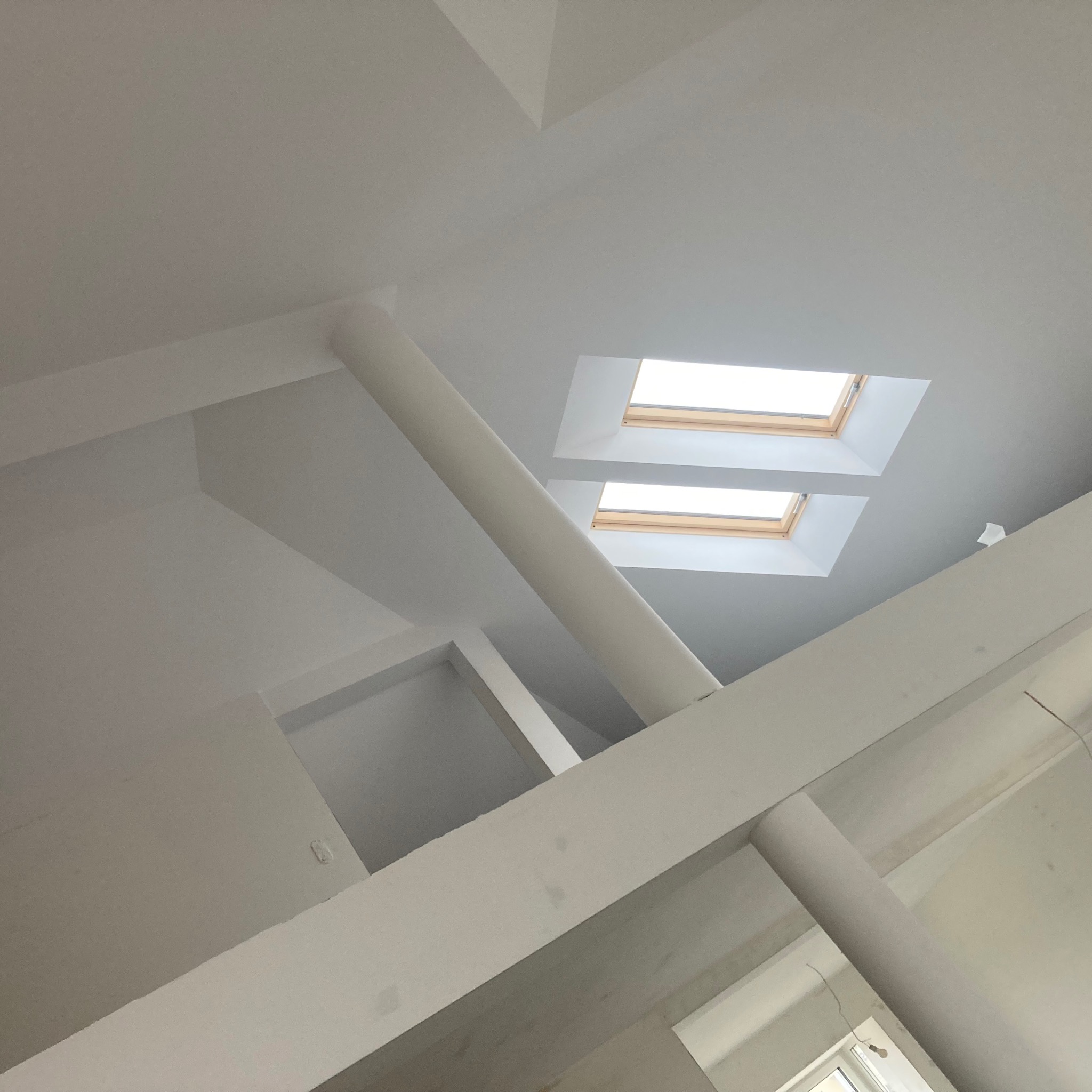 Białe, świeżo pomalowane poddasze z dwoma oknami dachowymi, widoczne belki konstrukcyjne i słup nośny, surowe wykończenie.