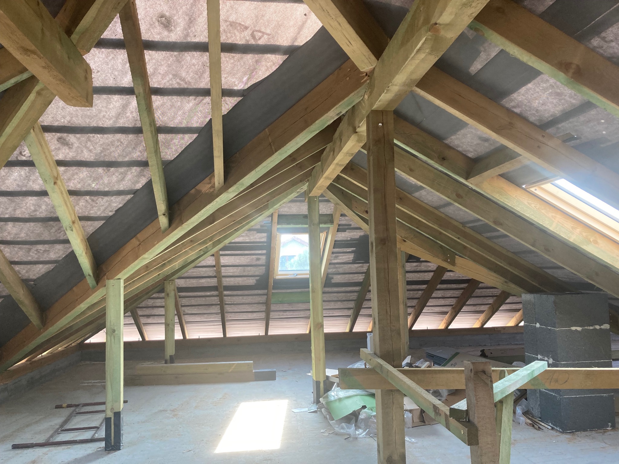 Konstrukcja drewniana poddasza z widoczną membraną dachową, oknem dachowym i kominem w trakcie budowy, częściowo oświetlona promieniami słonecznymi.