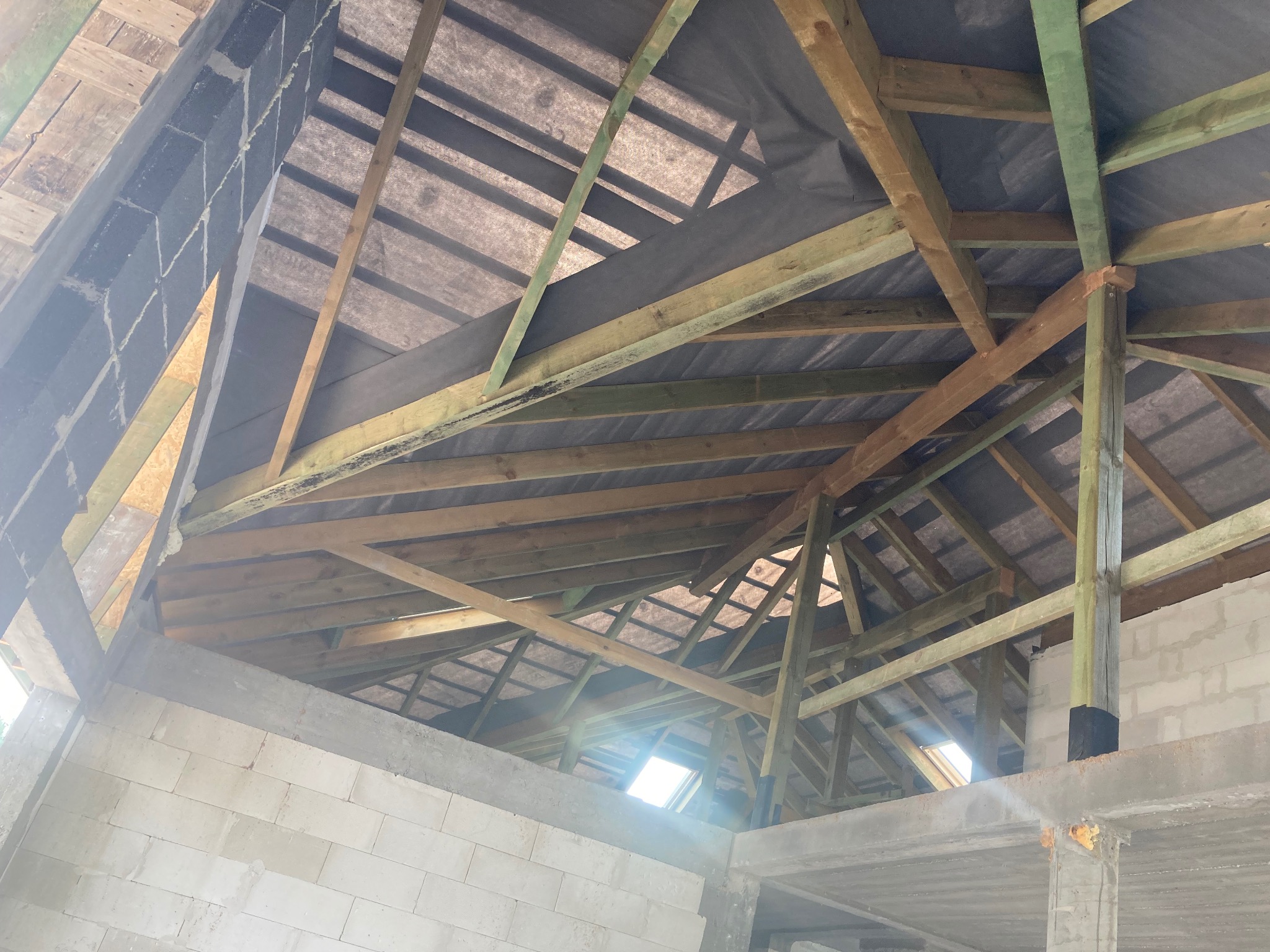 Szkielet dachu z drewnianych belek i czarnej membrany, widok od wewnątrz, w tle okno dachowe w surowym stanie.