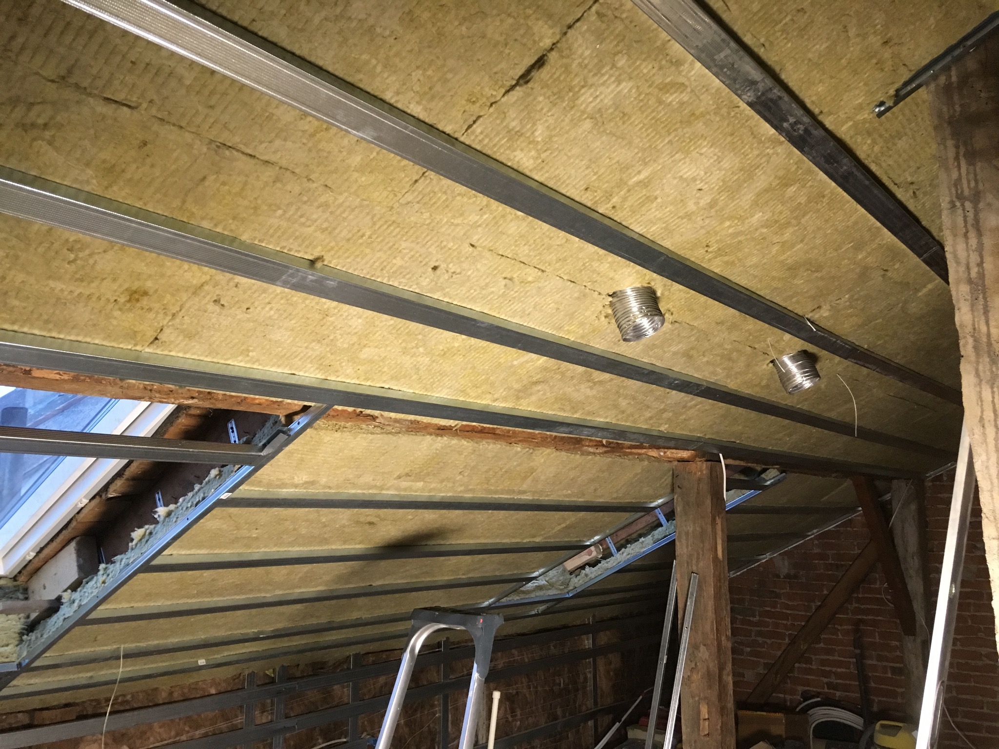 Izolacja termiczna poddasza z widocznymi metalowymi profilami, fragment okna dachowego i elementami wentylacji; widoczna drabina malarska.