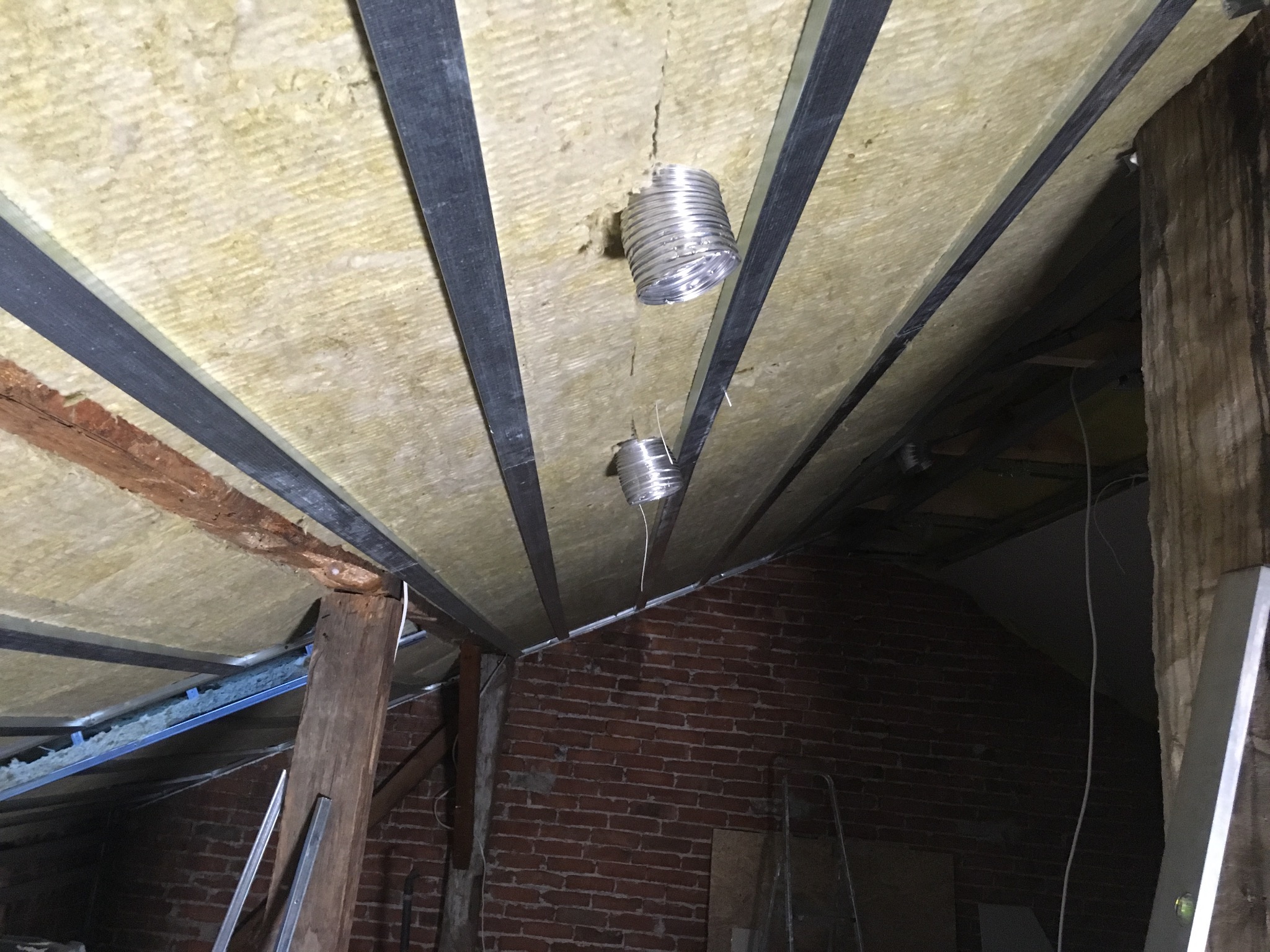 Ocieplenie poddasza wełną mineralną, widoczne profile metalowe, zawieszone elementy wentylacyjne i fragment ściany z czerwonej cegły.