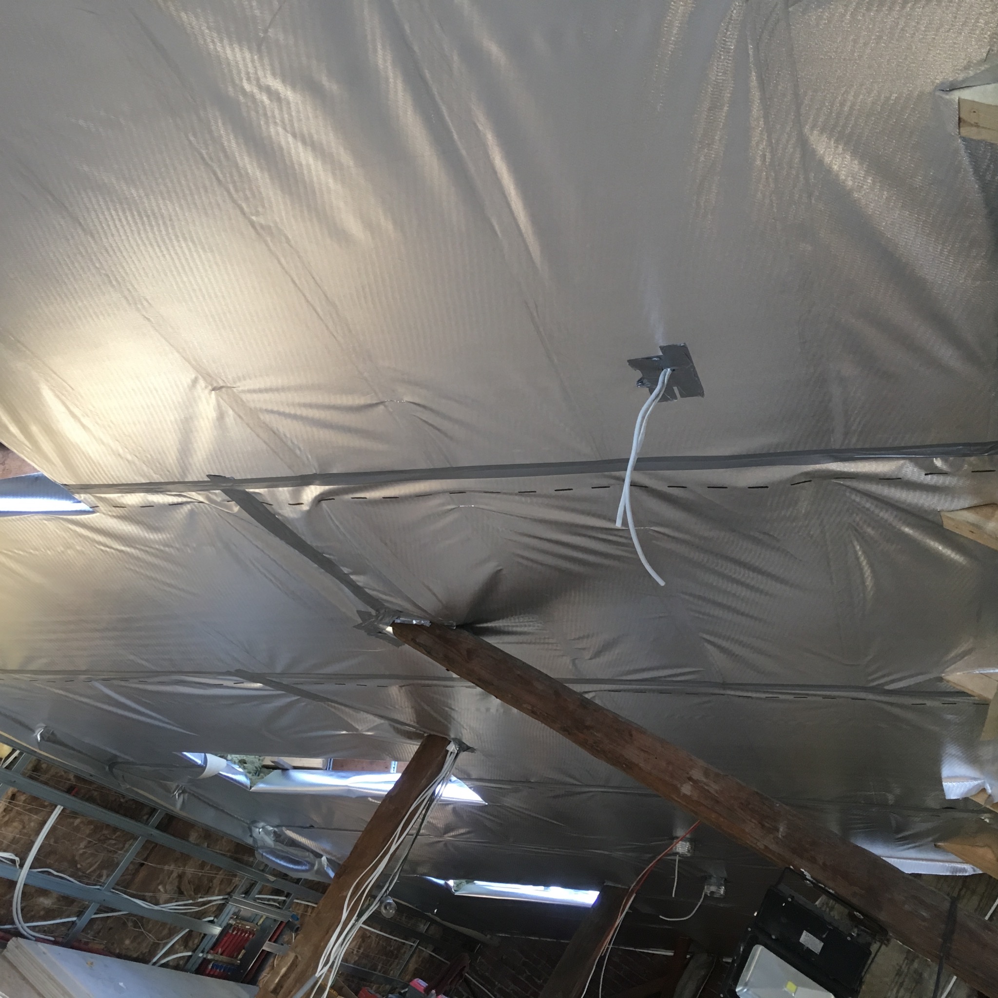 Srebrna folia termoizolacyjna zamontowana na poddaszu, widoczne drewniane belki konstrukcyjne i wystające przewody elektryczne z puszki.