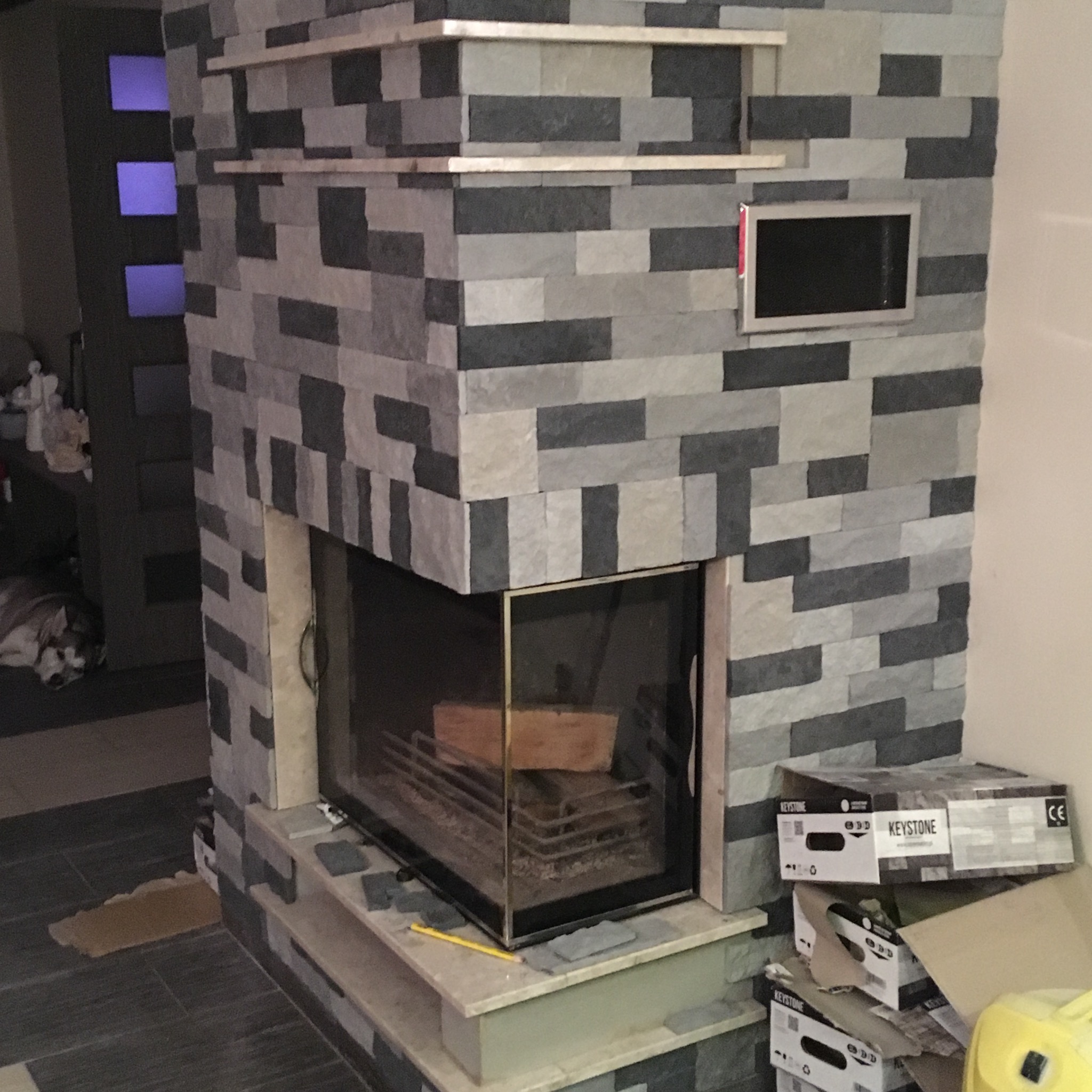 Wykończony kominek obłożony szarą, prostokątną płytką imitującą kamień, z wkładem kominkowym, drewnem wewnątrz i półkami na górze, widoczne kartony po płytkach.