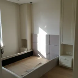 Rama łóżka tapicerowanego z wezgłowiem z paneli, wkomponowana między dwie wysokie szafy z wbudowanymi szafkami nocnymi, w jasnym wnętrzu.