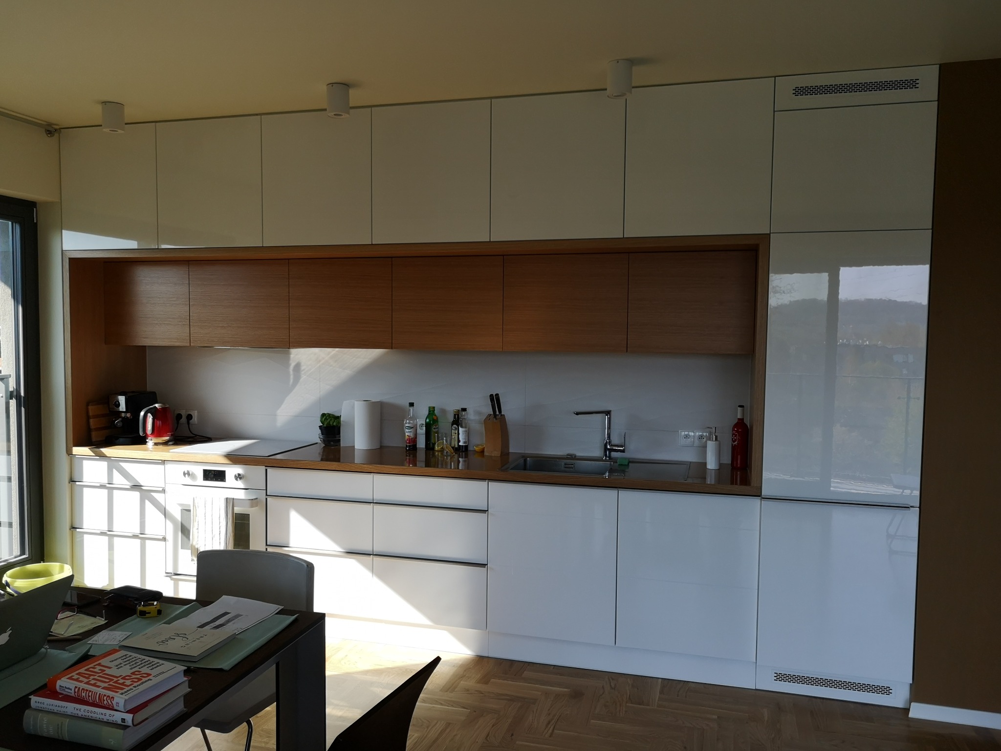 Jasna kuchnia z białymi, lakierowanymi frontami szafek i drewnianymi akcentami, oświetlona naturalnym światłem. Widoczny fragment blatu roboczego i stół z książkami.