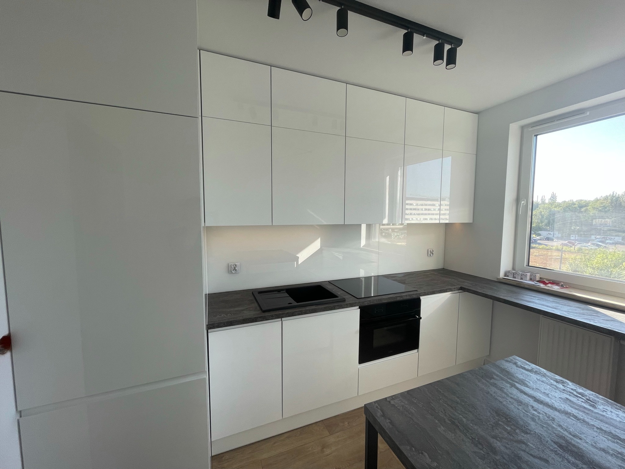 Nowoczesna, biała kuchnia z połyskiem, ciemny blat i oświetlenie punktowe. Ujęcie z bliska, widoczny fragment stołu i okno z widokiem na zewnątrz.