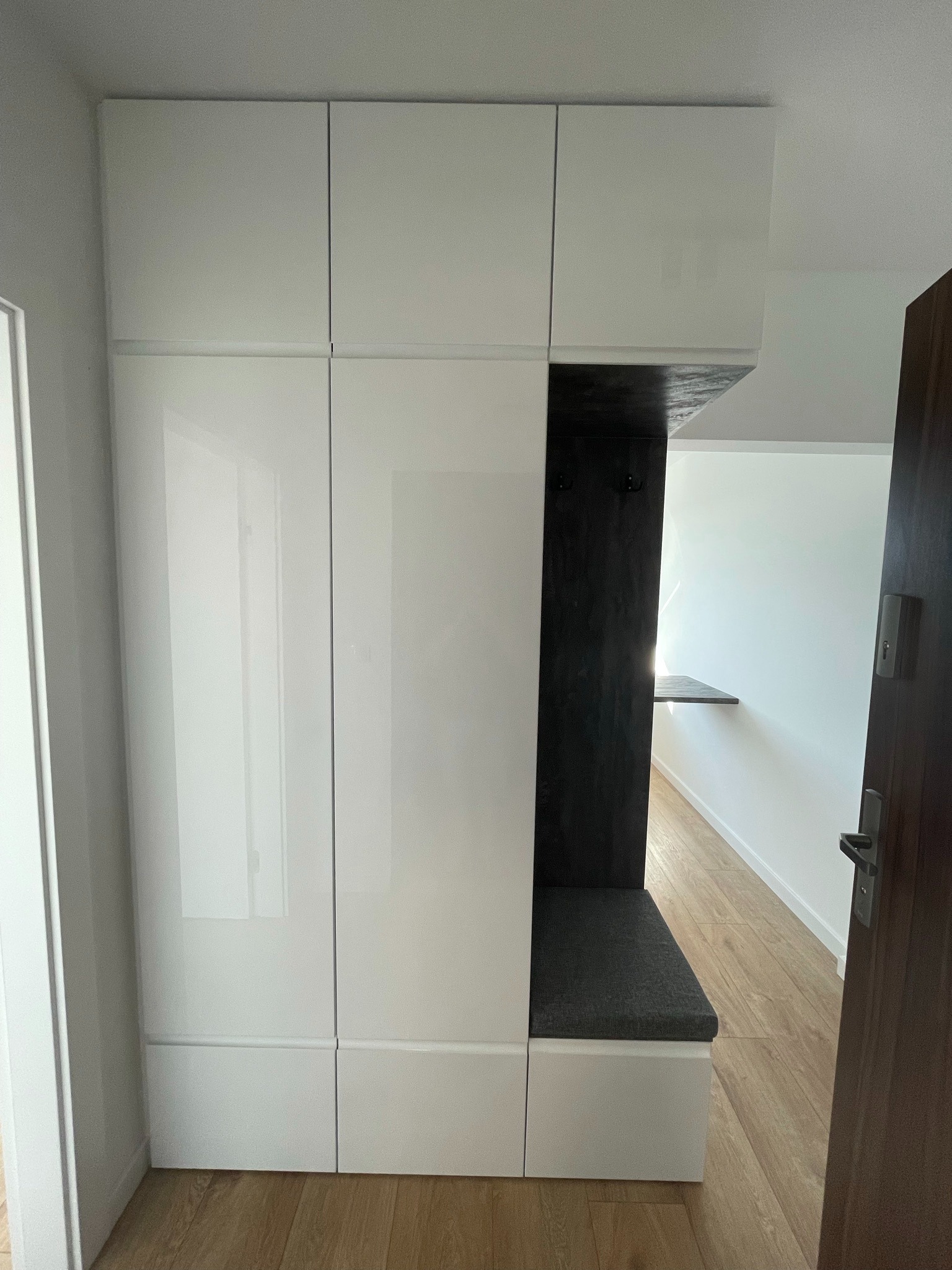 Biała szafa wnękowa z siedziskiem i wieszakami w przedpokoju, widok z perspektywy wejścia. Minimalistyczny design, jasne wnętrze, drewniana podłoga.