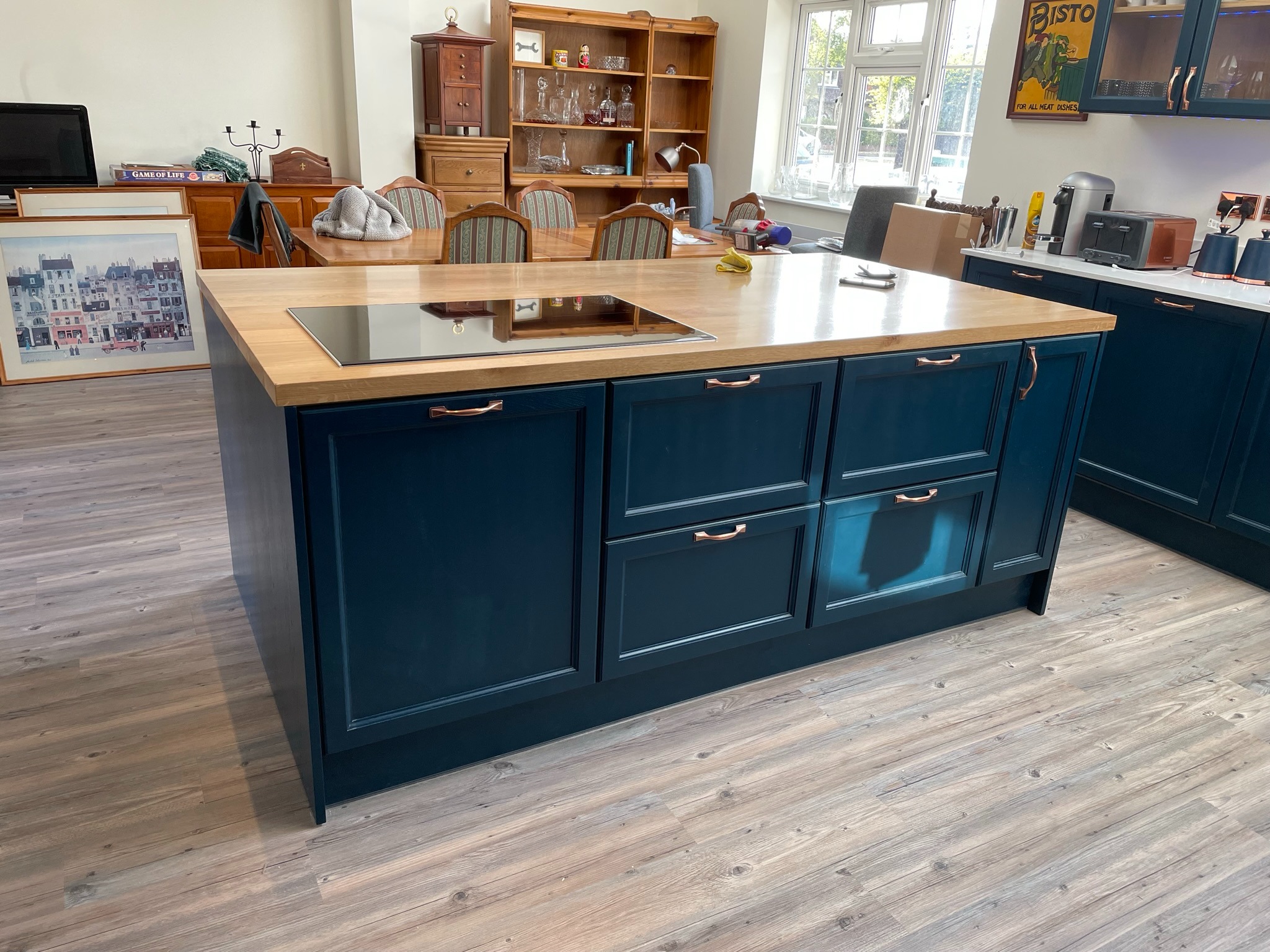 Wyspa kuchenna w odcieniach granatu z drewnianym blatem i płytą indukcyjną, miedziane uchwyty, na podłodze panele imitujące drewno. W tle stół i krzesła.