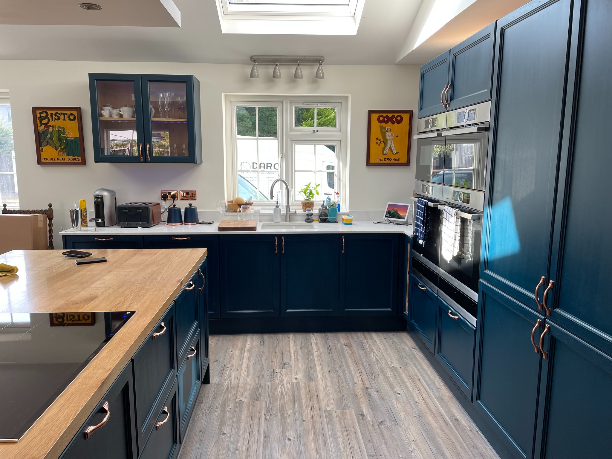 Niebieska kuchnia z drewnianym blatem i miedzianymi uchwytami. Widoczne sprzęty AGD, okno z widokiem na zewnątrz i plakaty na ścianie. Nowoczesny design i funkcjonalność.