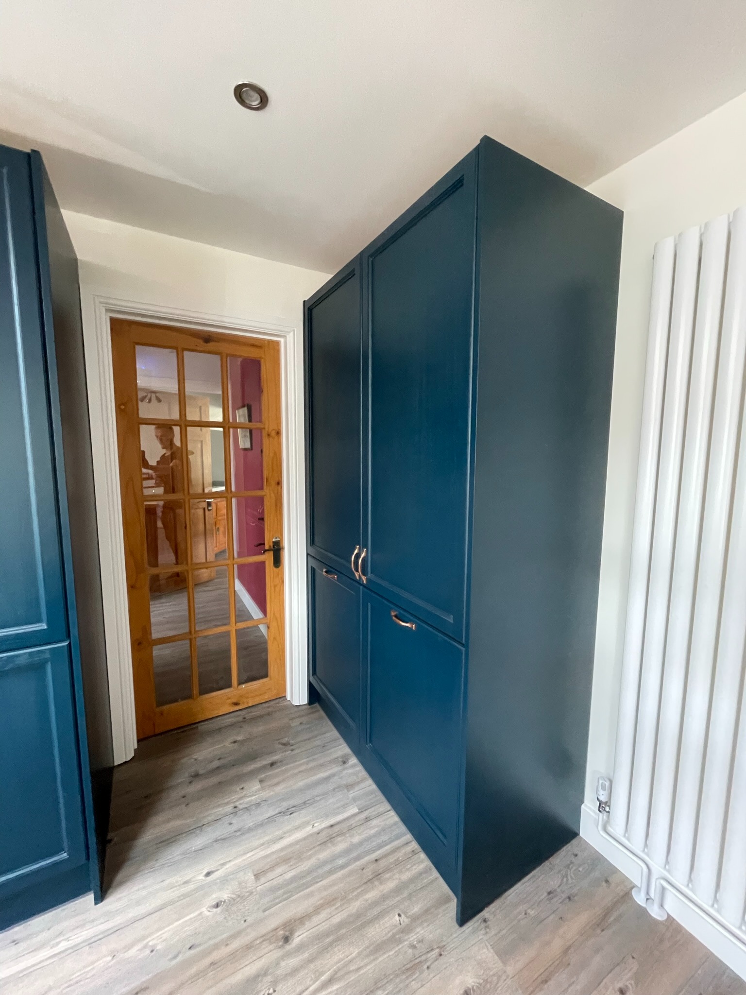 Granatowa szafa na wymiar z miedzianymi uchwytami, drewniana podłoga, biały grzejnik. Widoczne drzwi z szybami i odbicie osoby w środku.