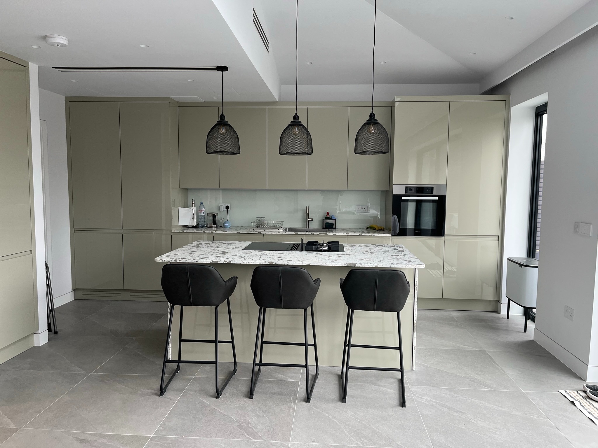 Nowoczesna kuchnia z wyspą i trzema hokerami. Fronty szafek w odcieniach beżu, marmurowy blat wyspy, trzy lampy wiszące z czarnymi kloszami.